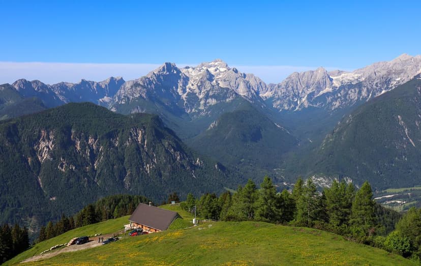 Dovška Baba mountain view with alpine meadow, wooden hut, and jagged limestone peaks under a clear blue sky.