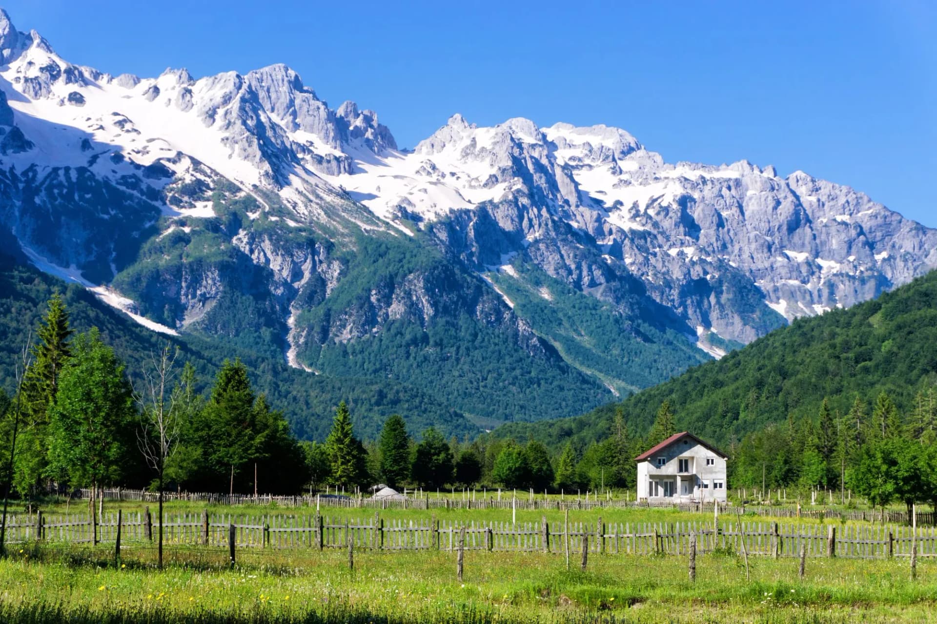 House in grassy field with wooden fence below snow-capped mountains in Valbona Village, Albania.
