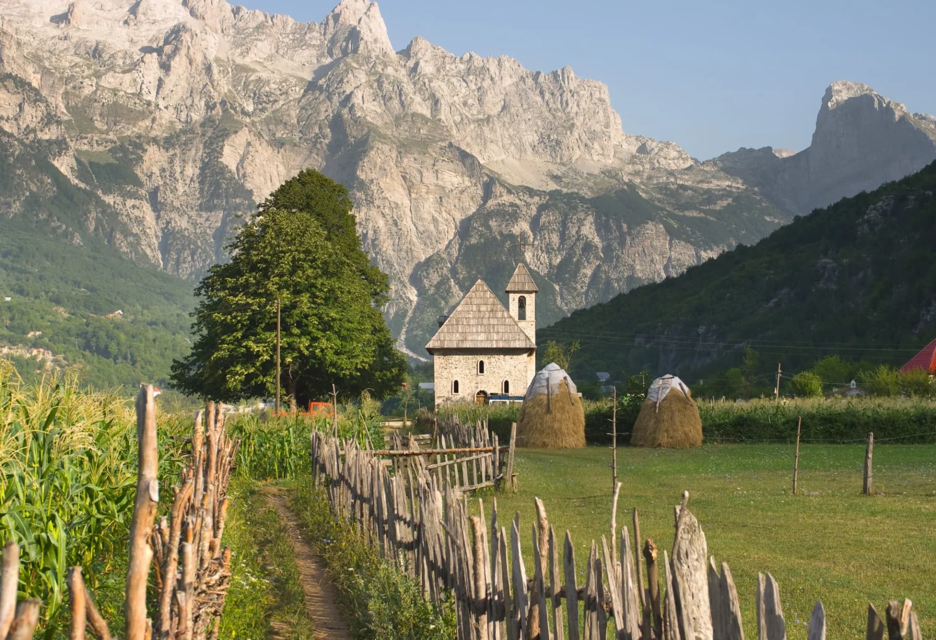 Stone church in Theth Valley, Albanian Alps, with haystacks and wooden fence in foreground.