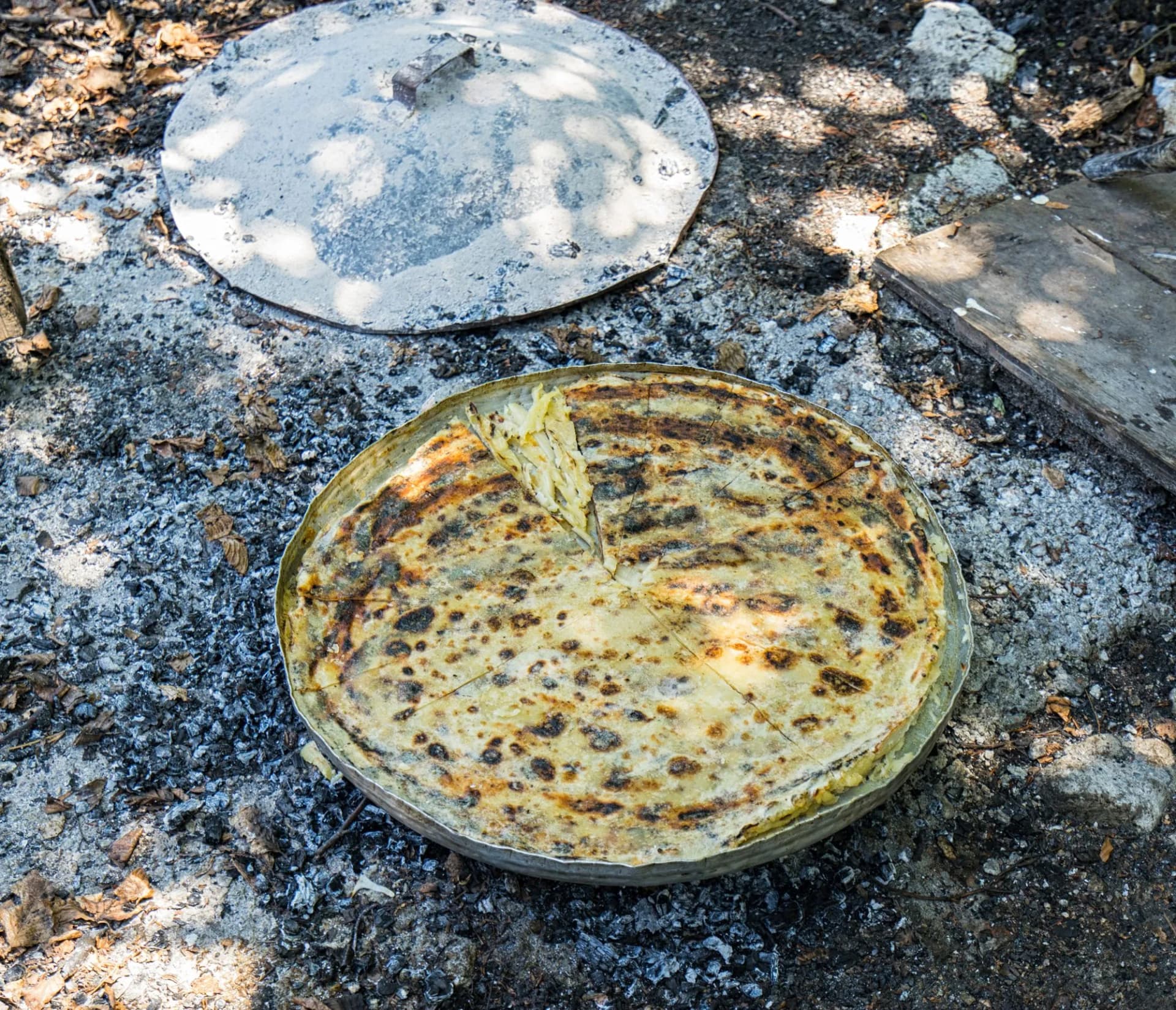 Traditional Albanian Flija pastry cooking outdoors over hot ashes with a metal lid nearby.