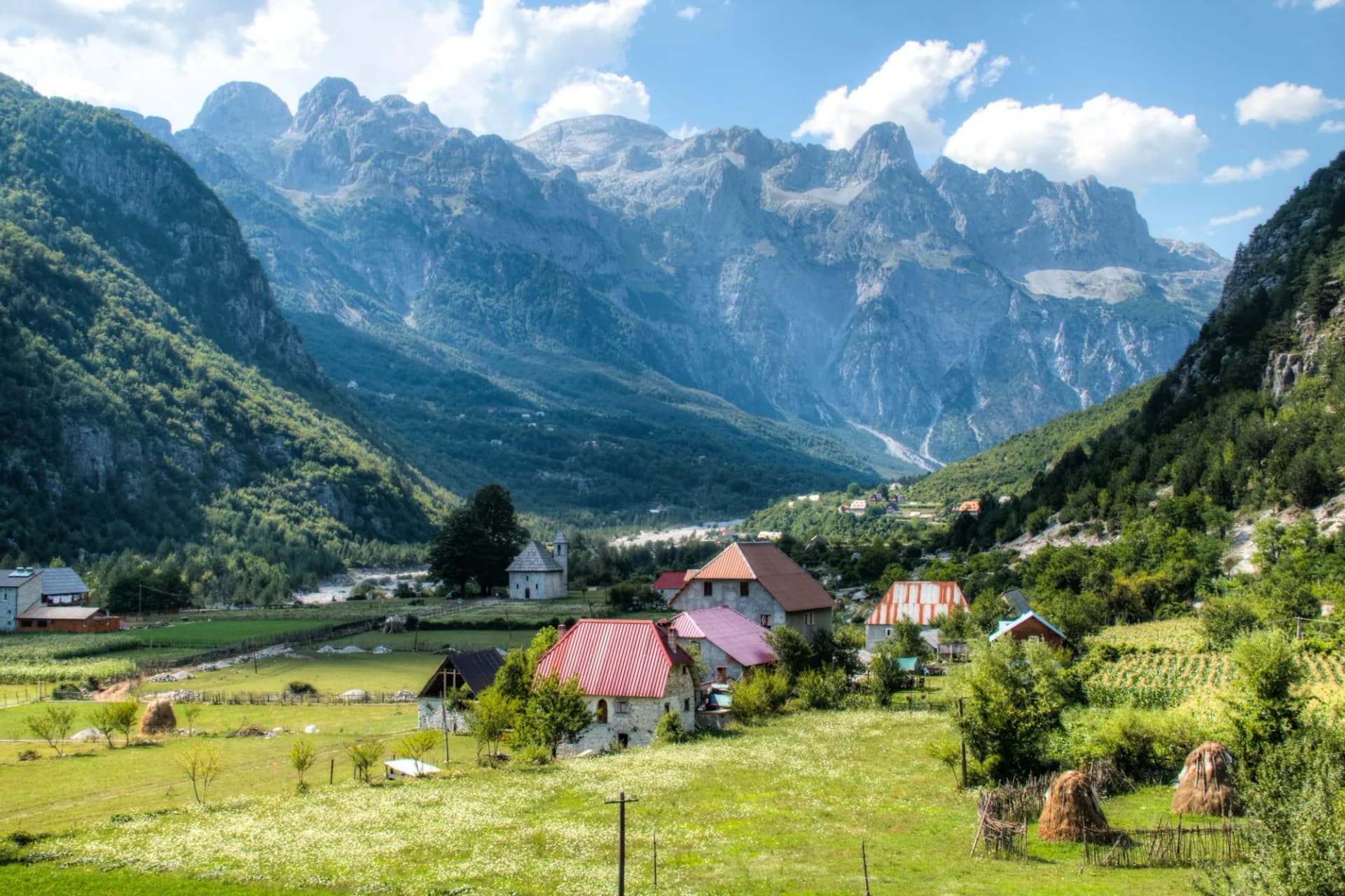 Mountain landscape in Theth with stone houses, green fields, and rugged peaks under a blue sky.
