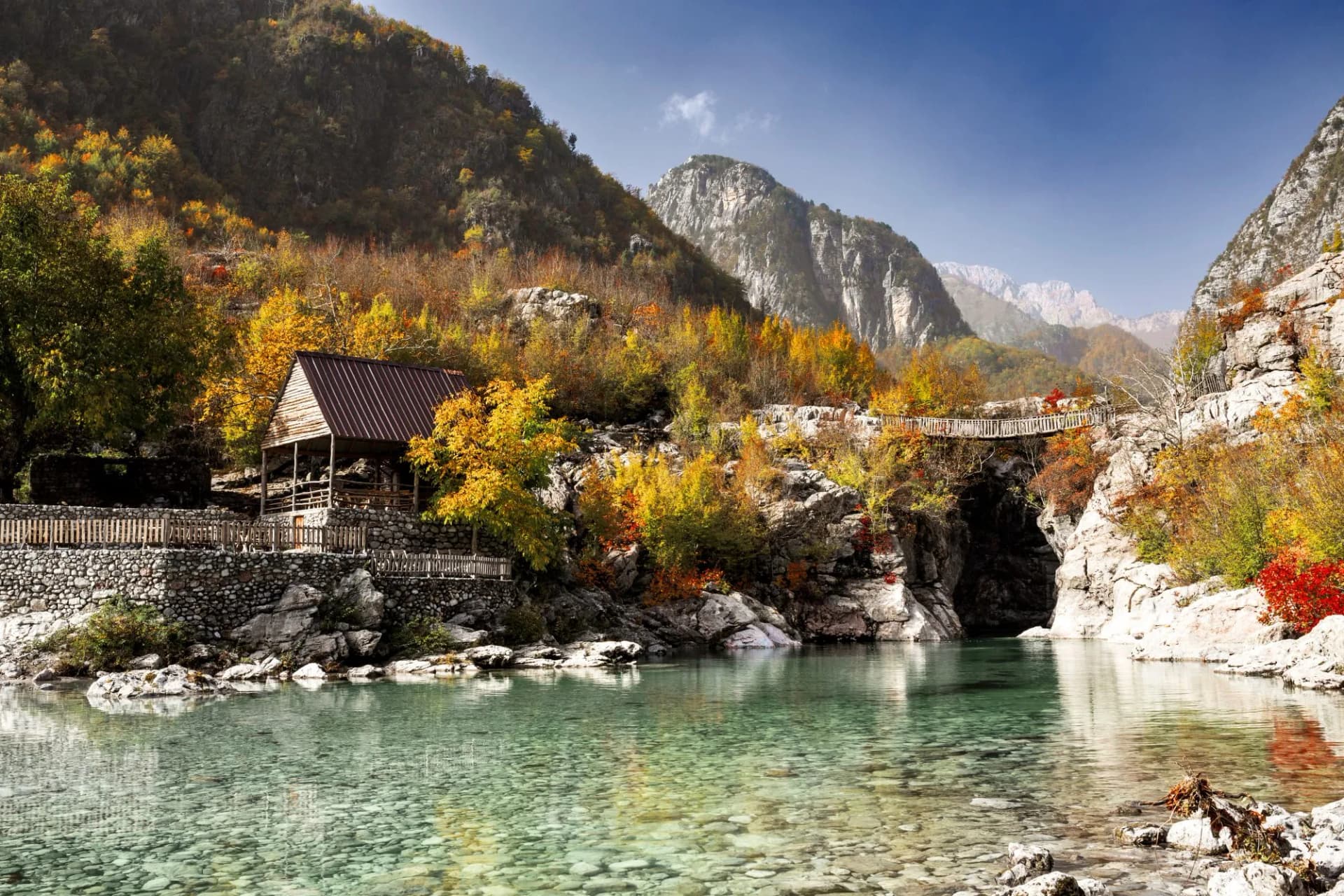 Clear river water flows past a wooden structure and footbridge nestled in mountains with autumn foliage.