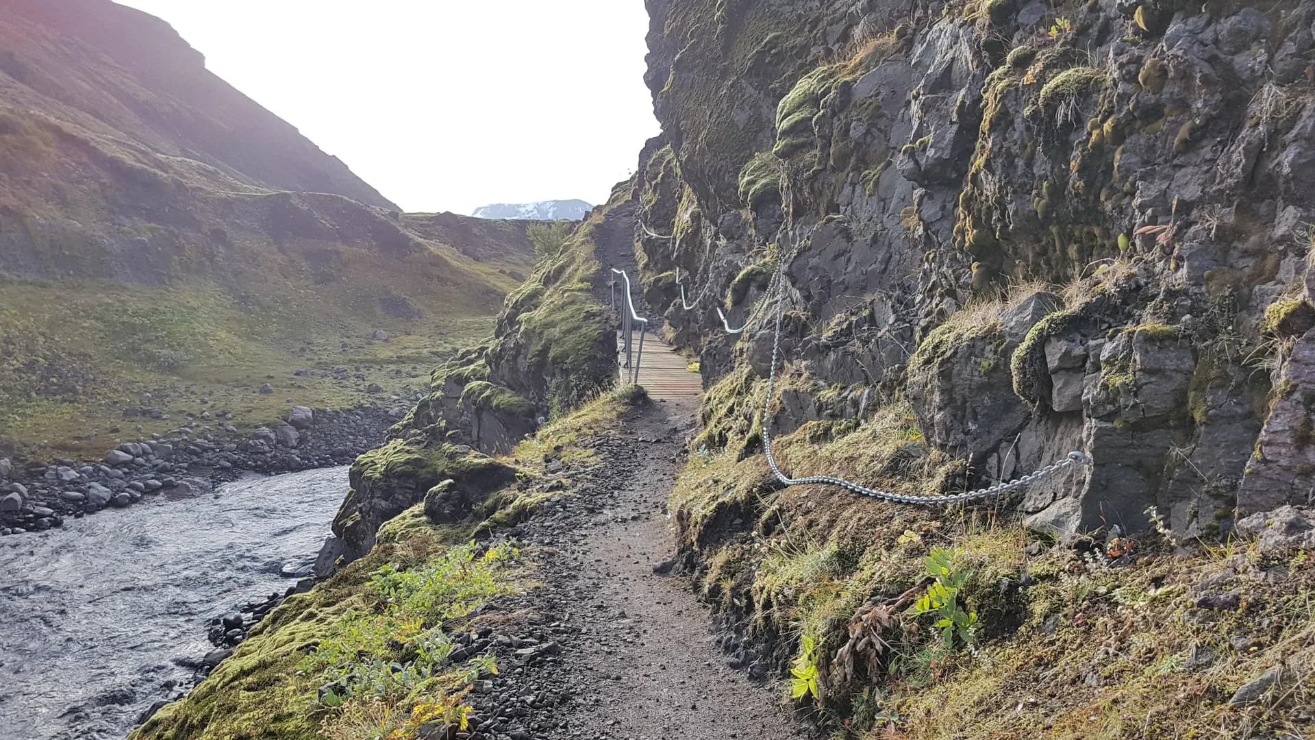 Hiking trail with metal safety chain along mossy rock face and river on the Laugavegur trail in Iceland.