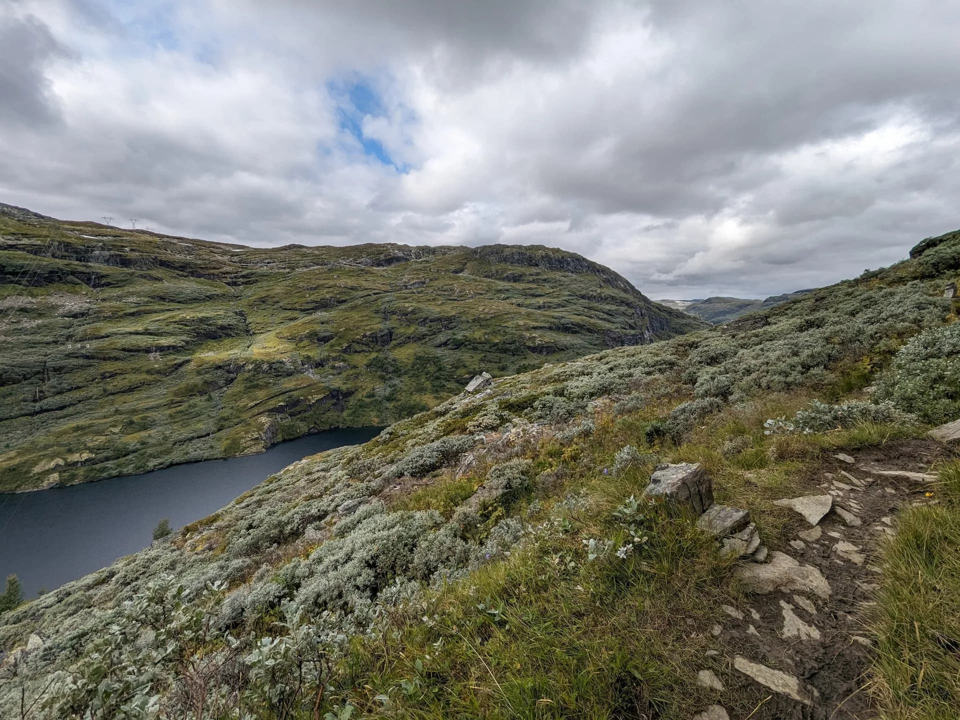 Hiking trail along a mountain lake in Aurlandsdalen, Norway, with green hills under a cloudy sky.