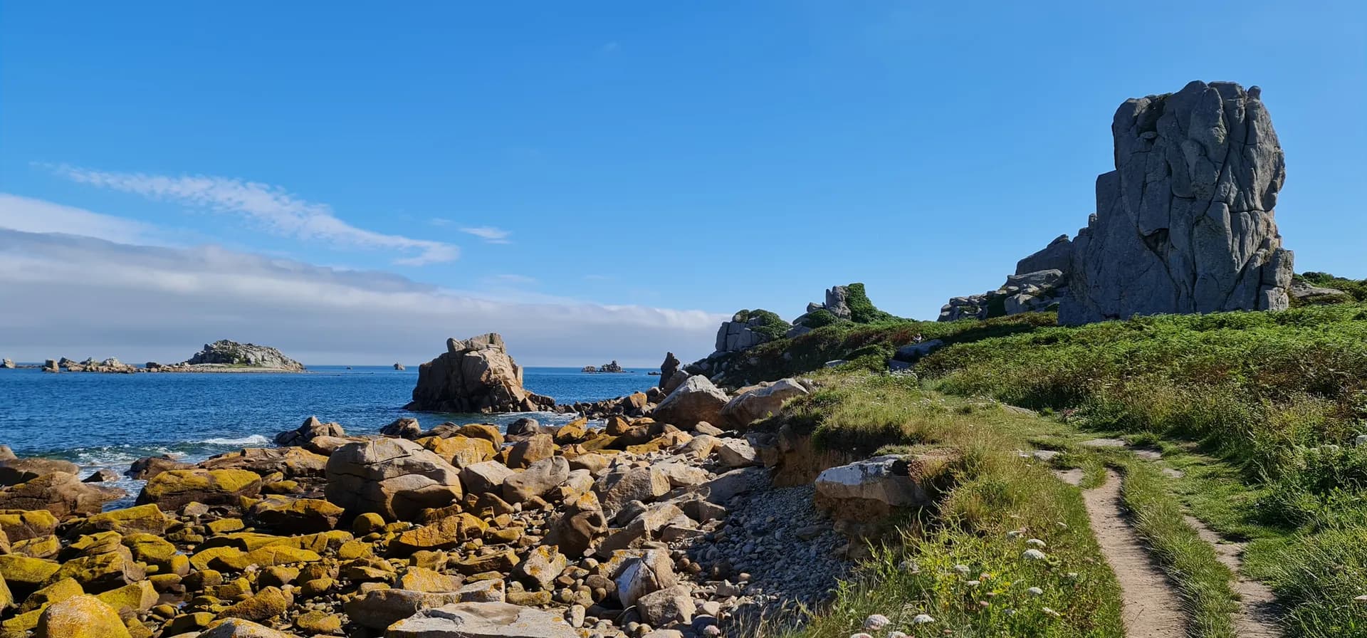 Rocky coastline with a dirt hiking path, large granite boulders, and blue ocean in Brittany, France.