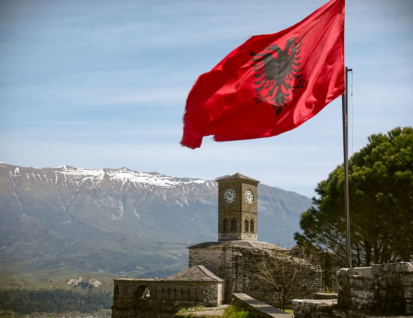 Albanian flag waving over a stone clock tower with snow-capped mountains in the background.