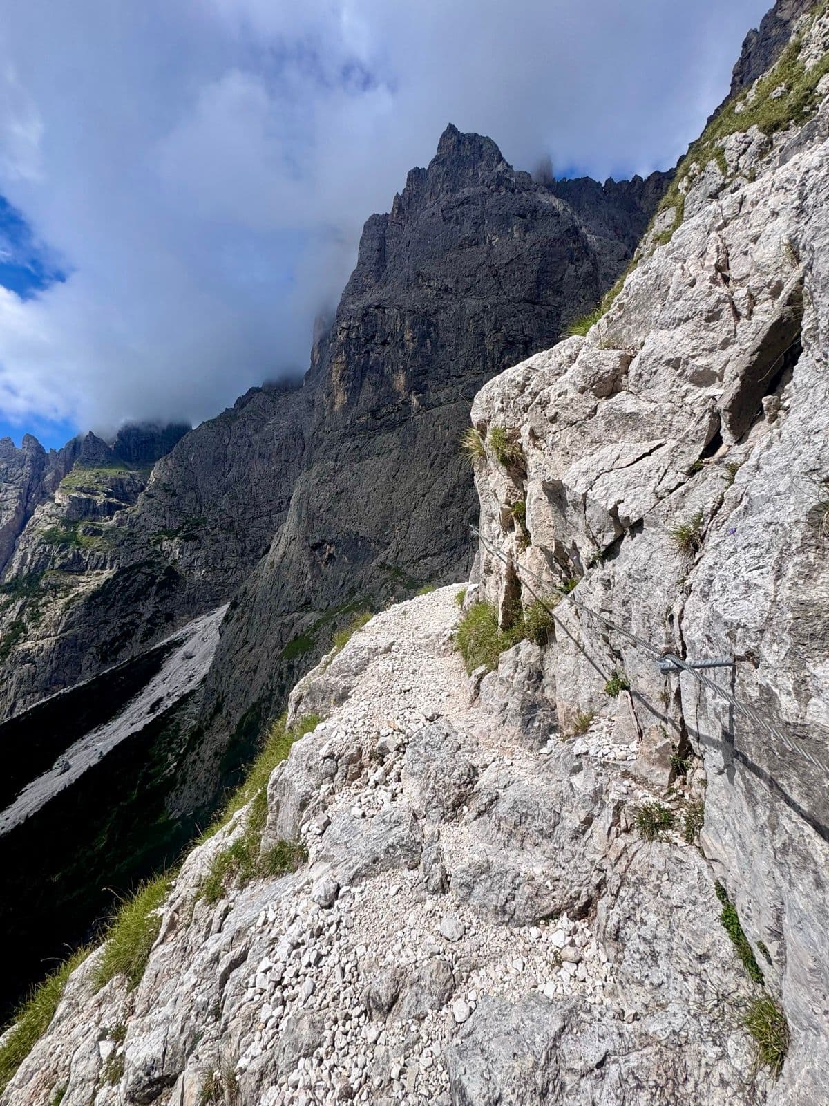 Narrow hiking trail with safety cable along a rocky cliff in the Pale di San Martino mountains.