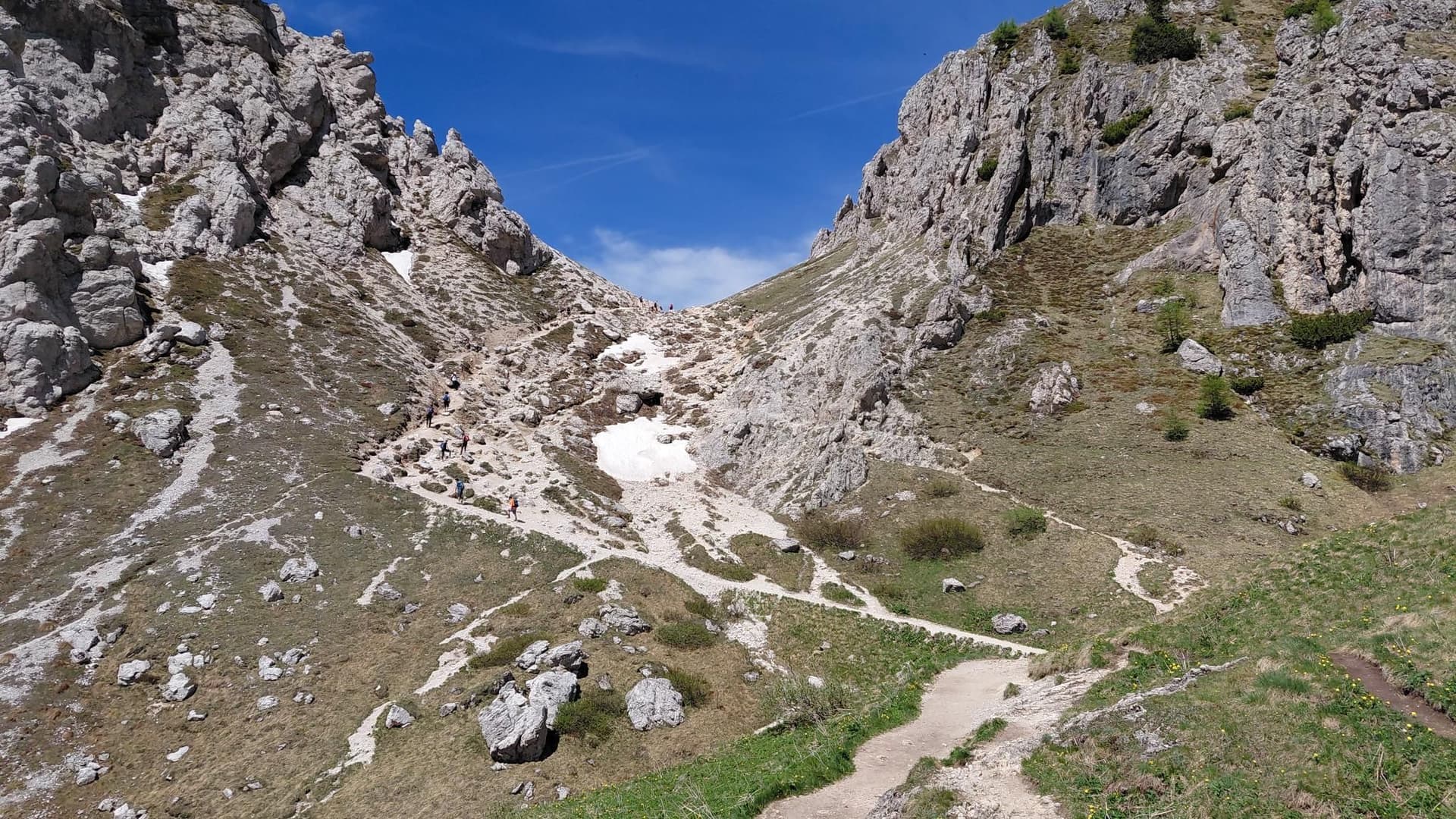 Hikers on a rocky mountain trail with patches of snow and steep limestone cliffs under a clear blue sky.