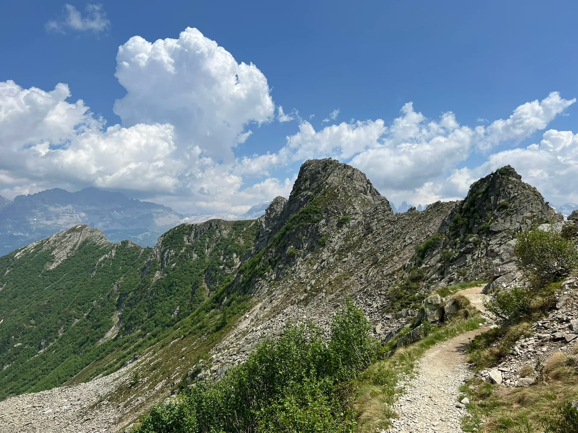 Hiking trail along rocky mountain ridges in Adamello-Brenta Nature Park with green slopes and blue sky.