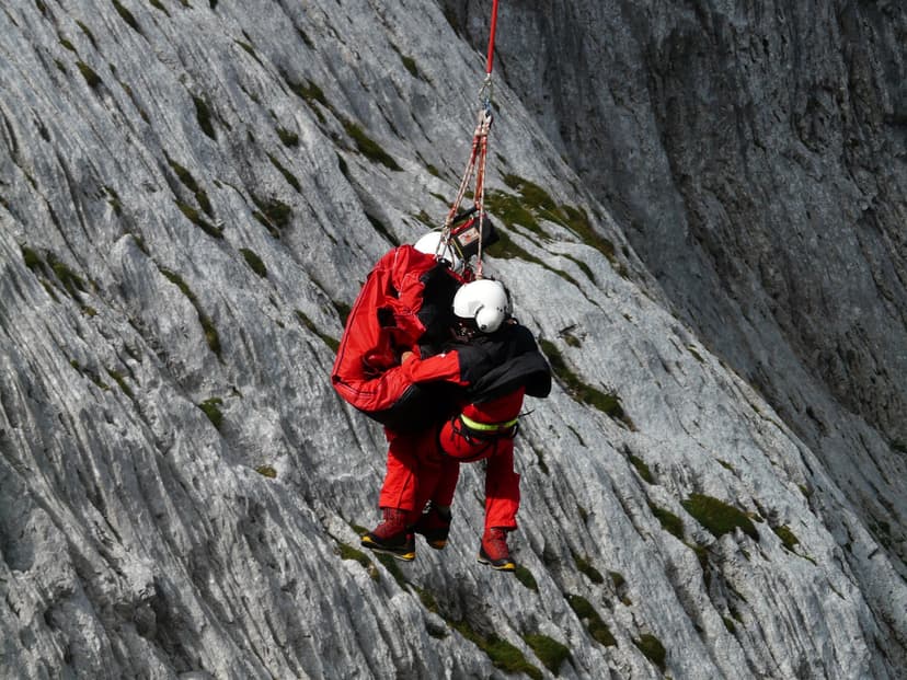 Rescue workers in red suits suspended from a helicopter hoist against a rocky mountain cliff face