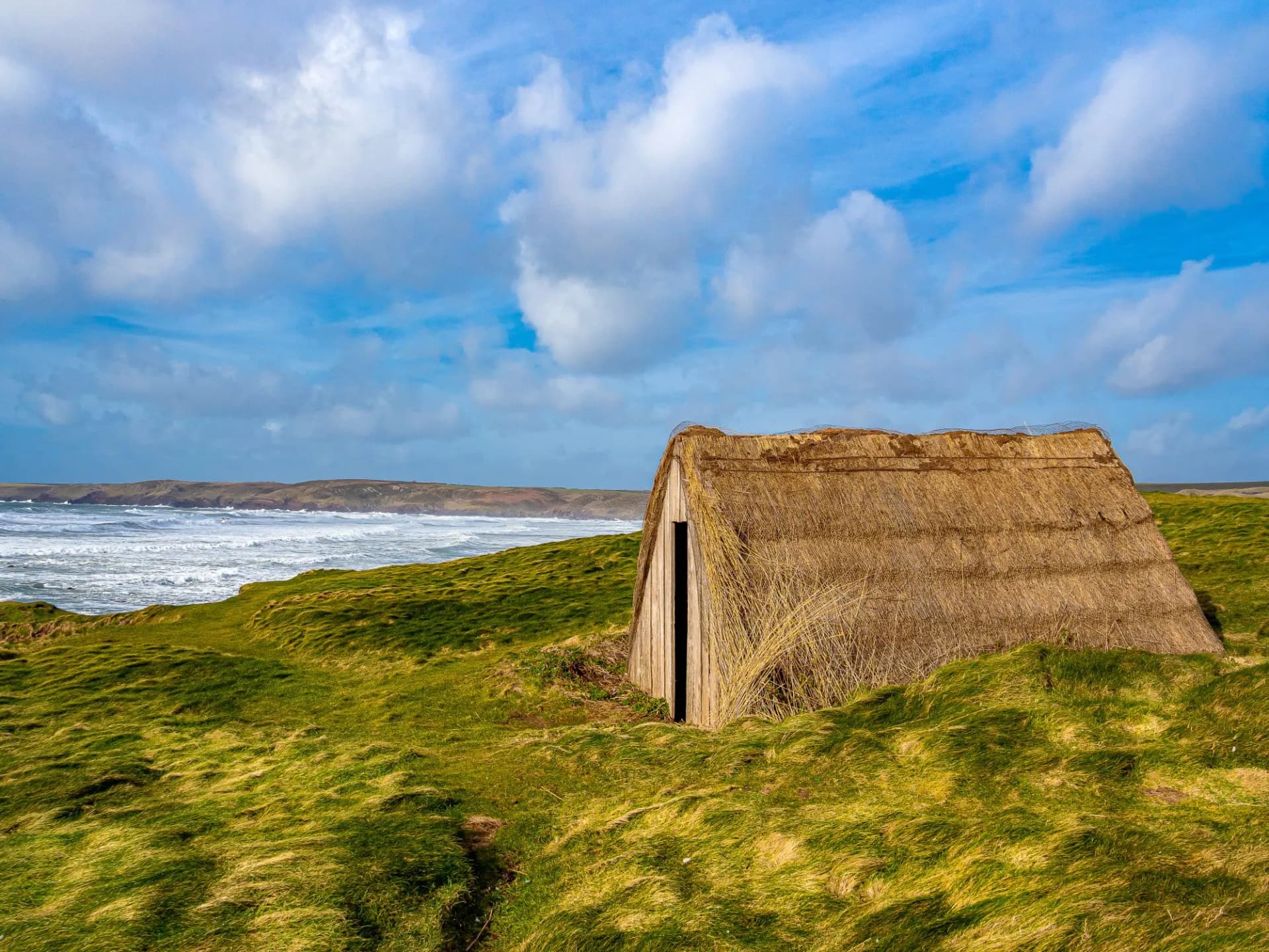 Thatched hut on grassy coastal dunes overlooking the rough sea under a blue, cloudy sky.