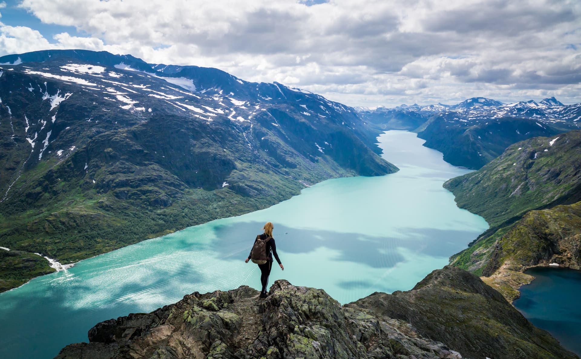 Hiker overlooking turquoise glacial lake surrounded by mountains with snow patches in Norway.