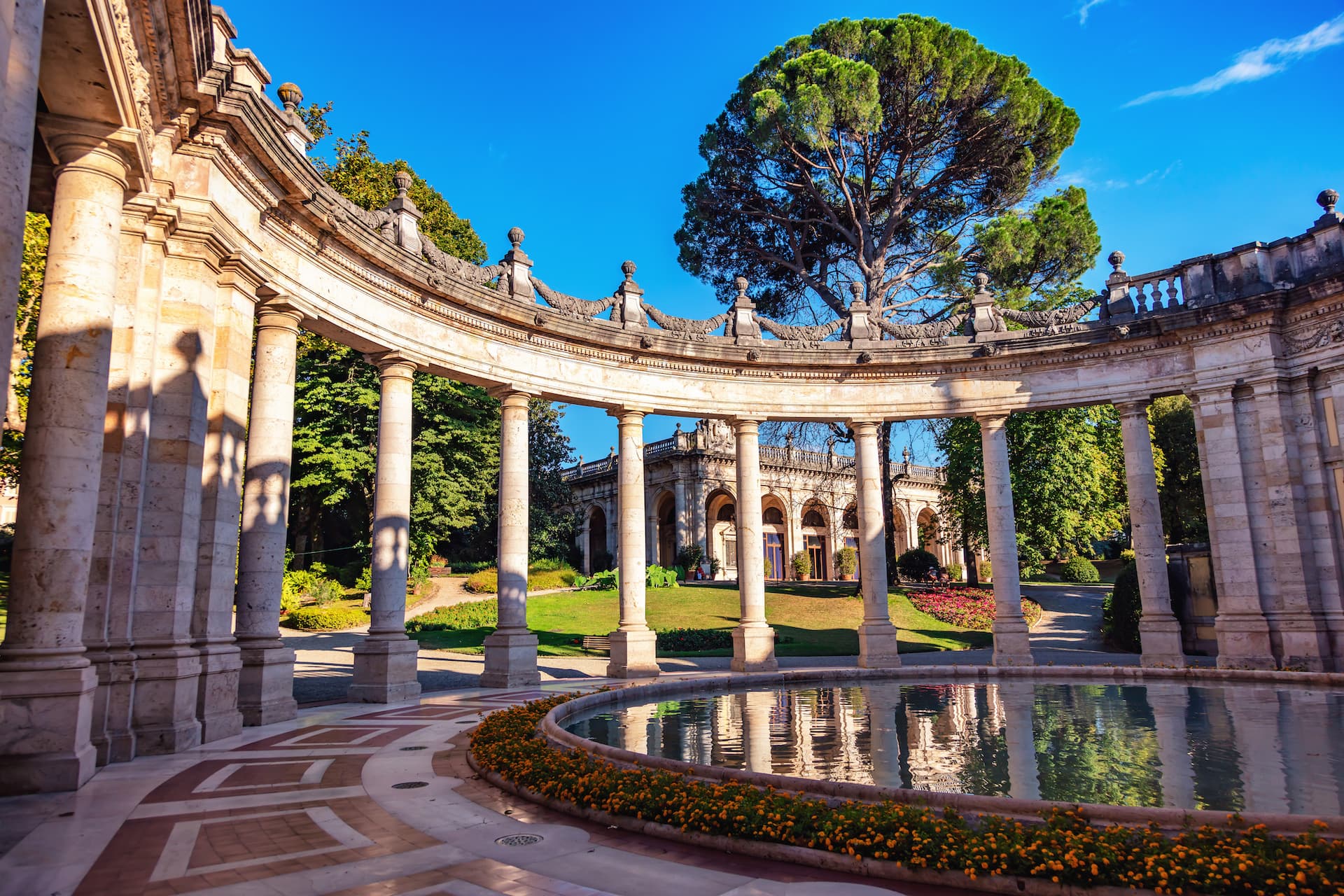 Curved colonnade framing a fountain and historic building with a large pine tree under blue sky.