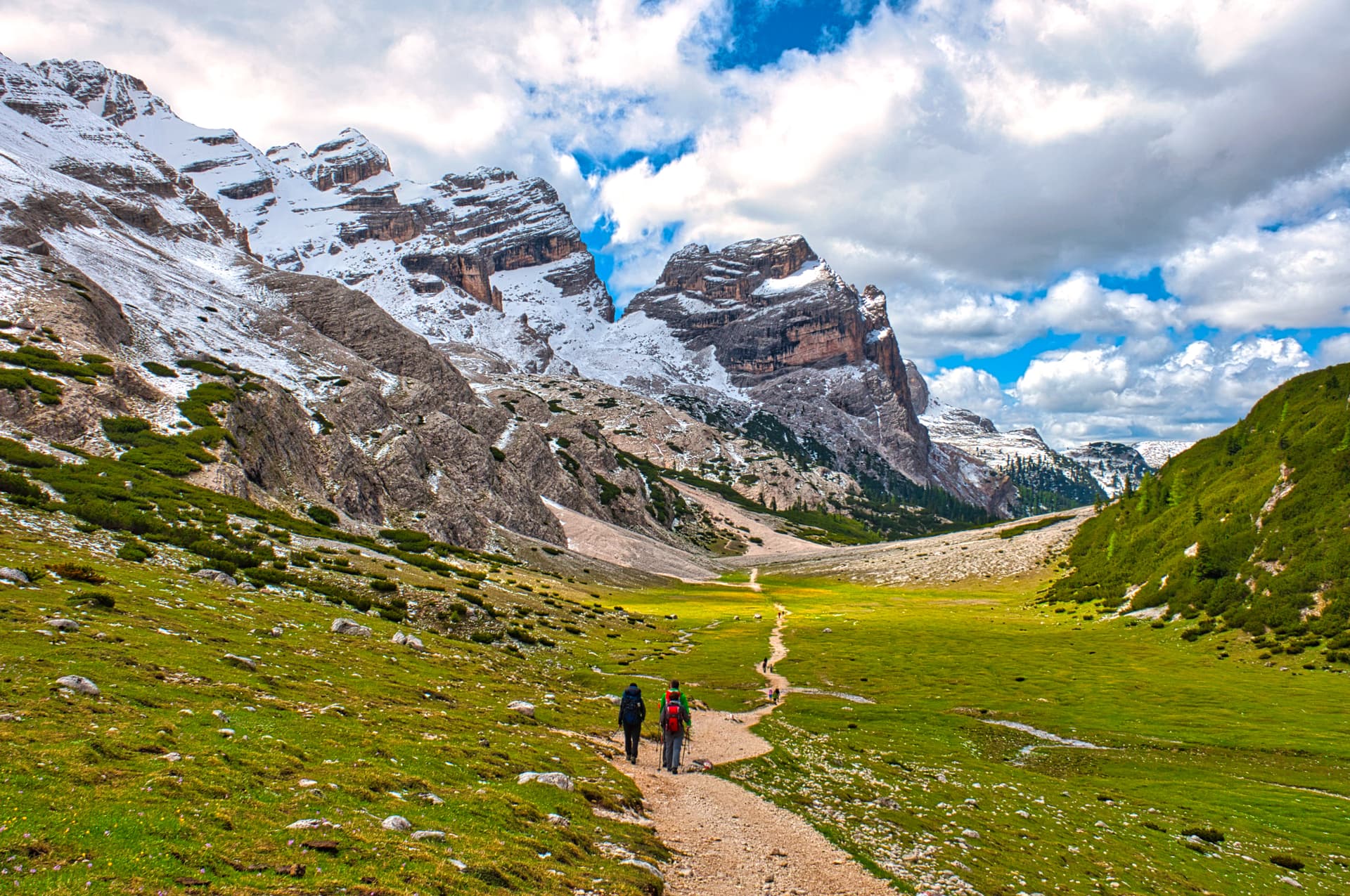 Hikers on dirt path through green valley toward snow-dusted mountains under blue sky.