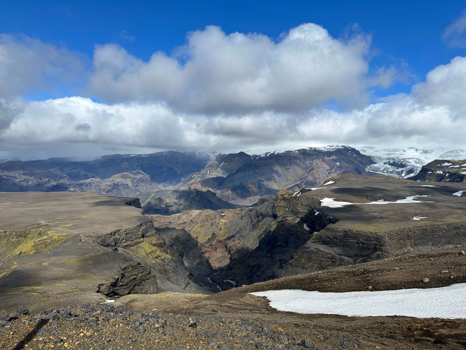 Volcanic plateau with deep canyon, snow patches, and glacier-capped mountains under blue sky.