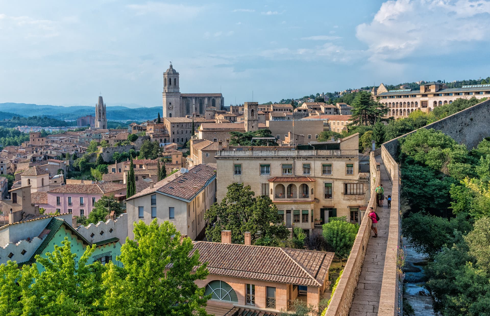 View of Girona city rooftops, cathedral, and ancient wall walkway with people walking.