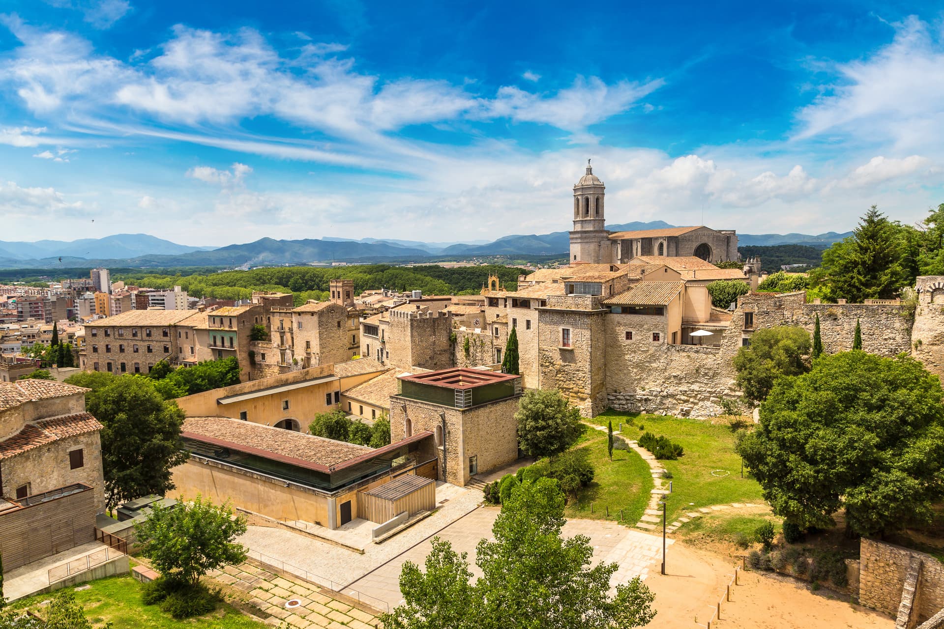 Aerial view of historic stone buildings and cathedral tower in Girona under a bright blue, cloudy sky.