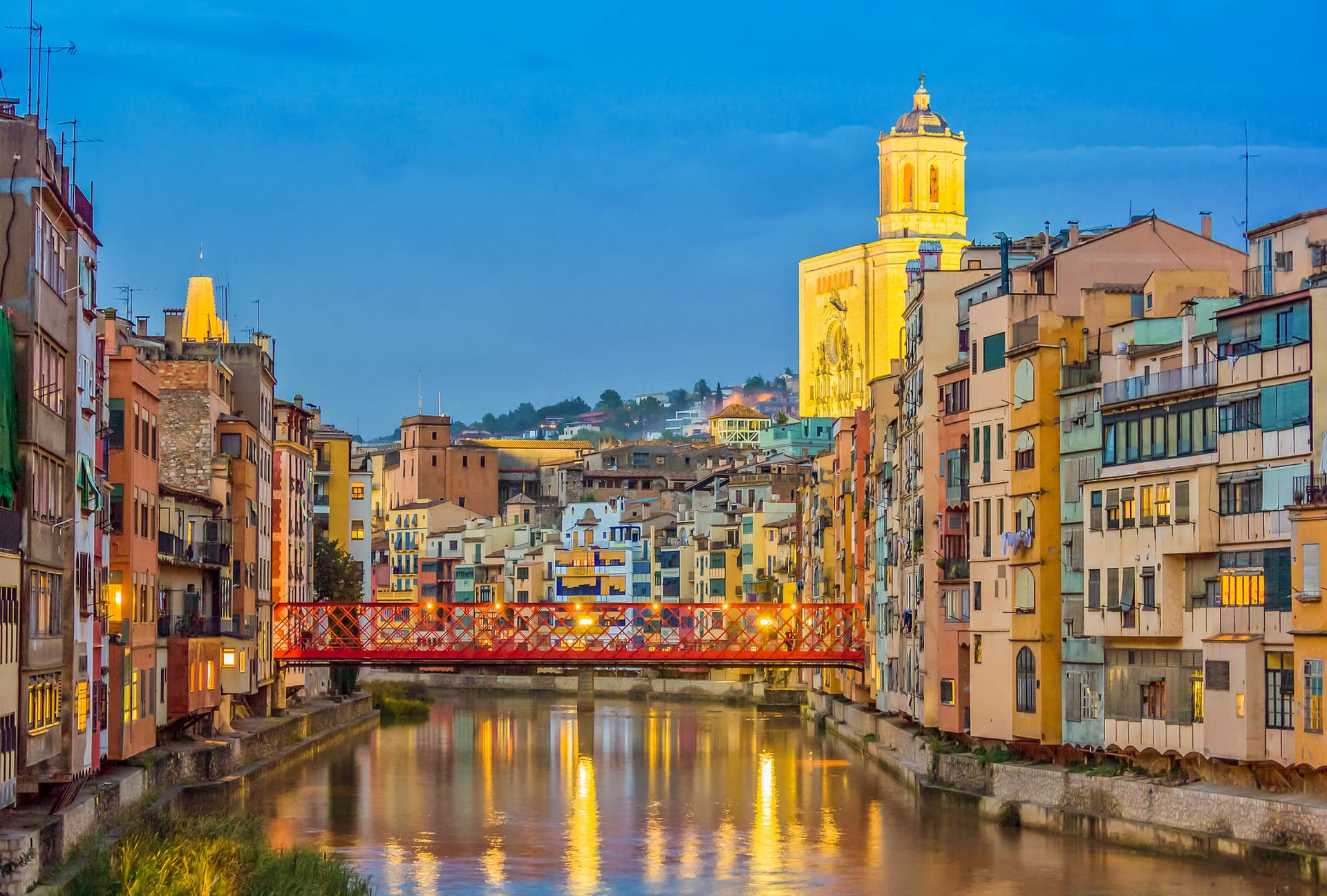 Colorful buildings line the river in Girona with a red bridge and illuminated cathedral tower at dusk.
