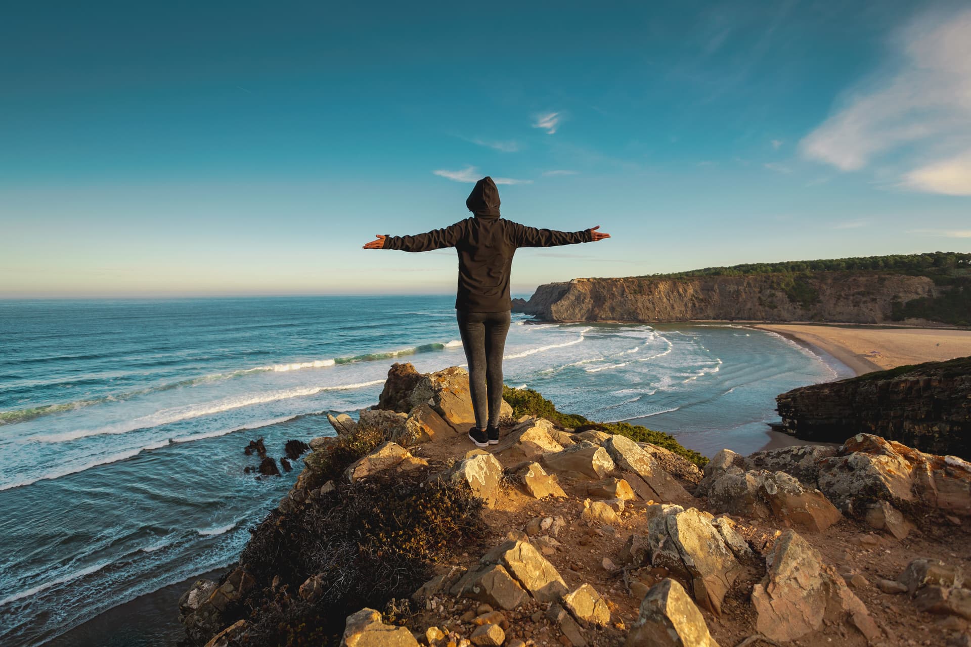 Hiker with arms outstretched on cliff edge overlooking beach and waves on Fishermen's Trail in Algarve, Portugal.