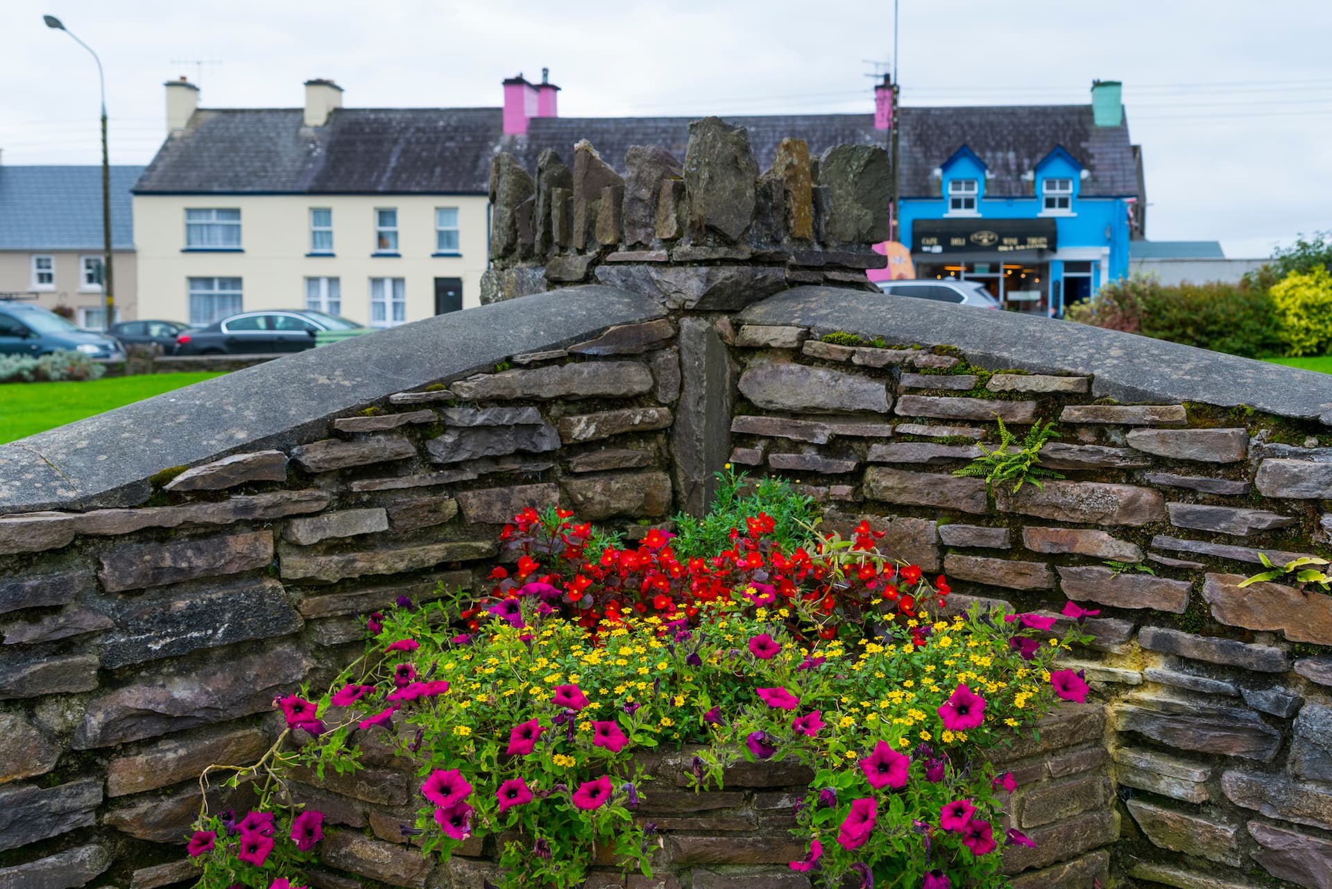 Stone wall planter with red, yellow, and purple flowers in Sneem Village with colorful buildings.