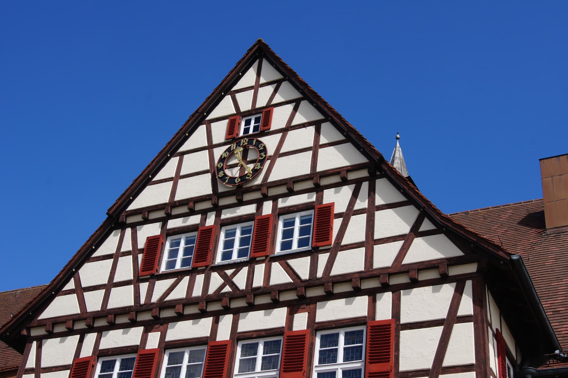 Half-timbered house gable with clock and red shutters against bright blue sky in Münsingen.