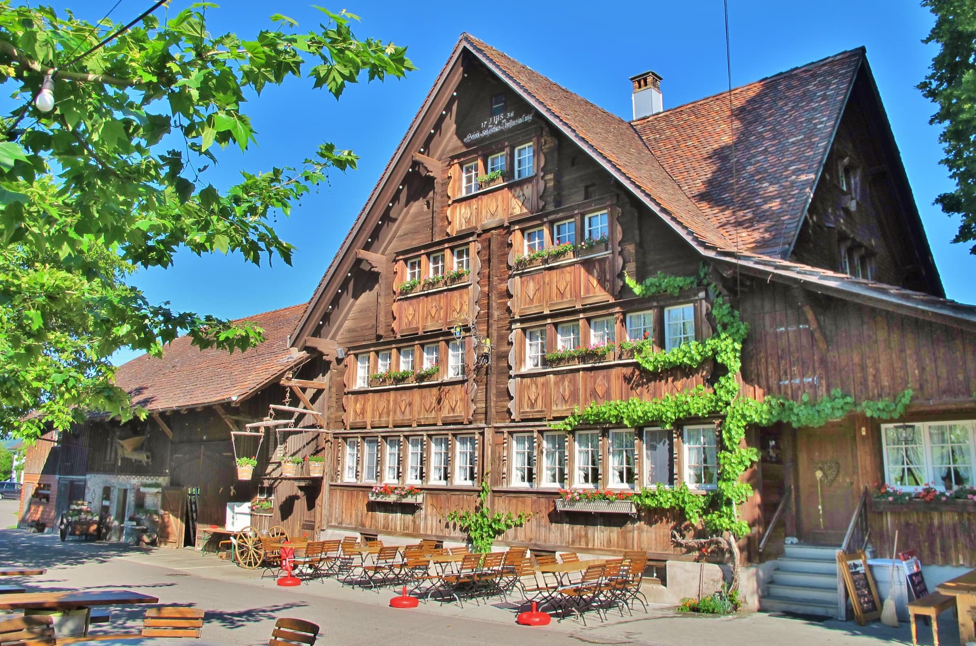 Traditional wooden Appenzell house with outdoor seating under green foliage and blue sky.