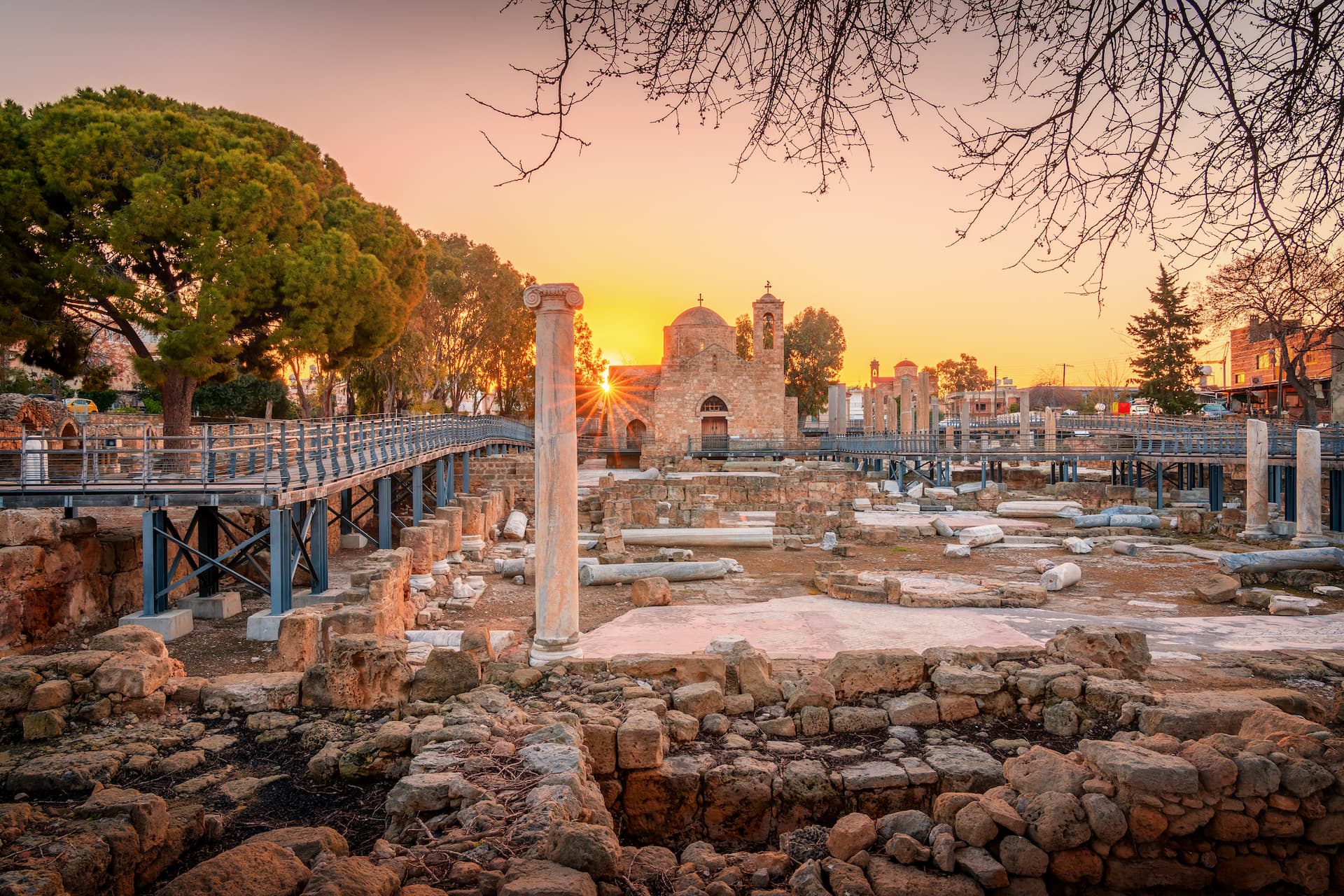 Archaeological ruins with a small church at sunset, viewed from behind a modern walkway in Paphos.