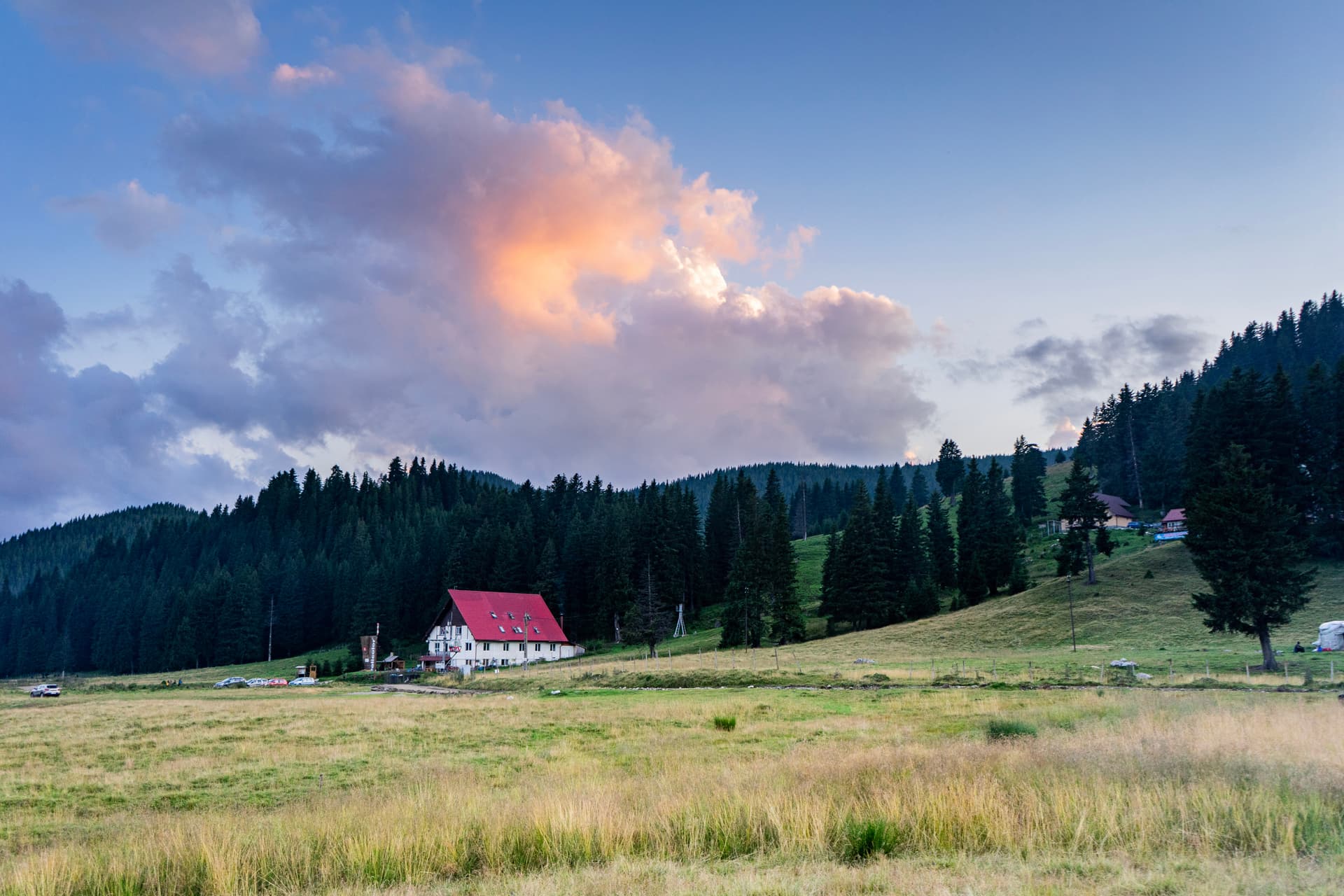 White mountain lodge with red roof nestled in grassy field below dark pine forest at sunset.