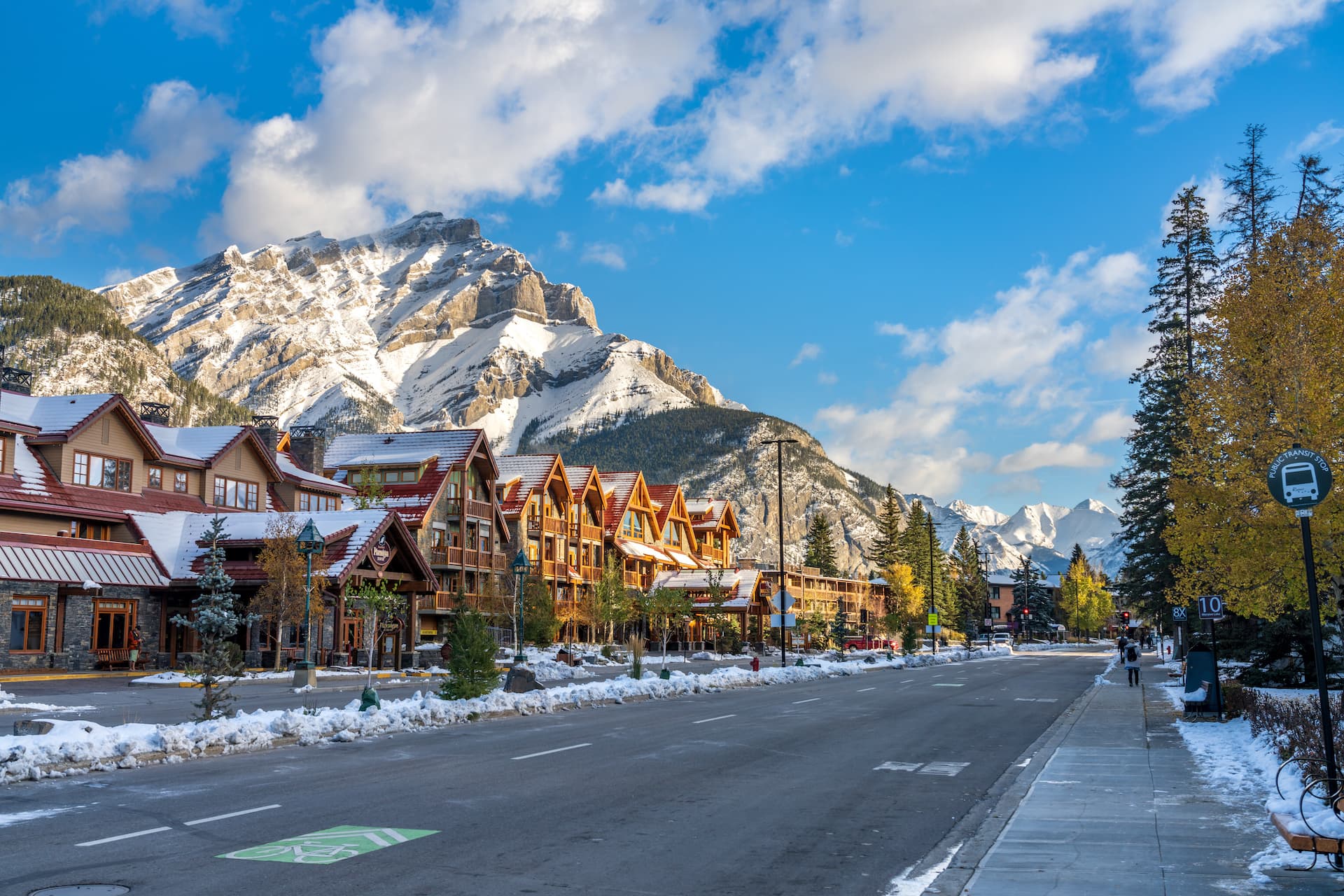 Street in Banff with snow-dusted buildings and large snow-capped mountains under a blue sky.