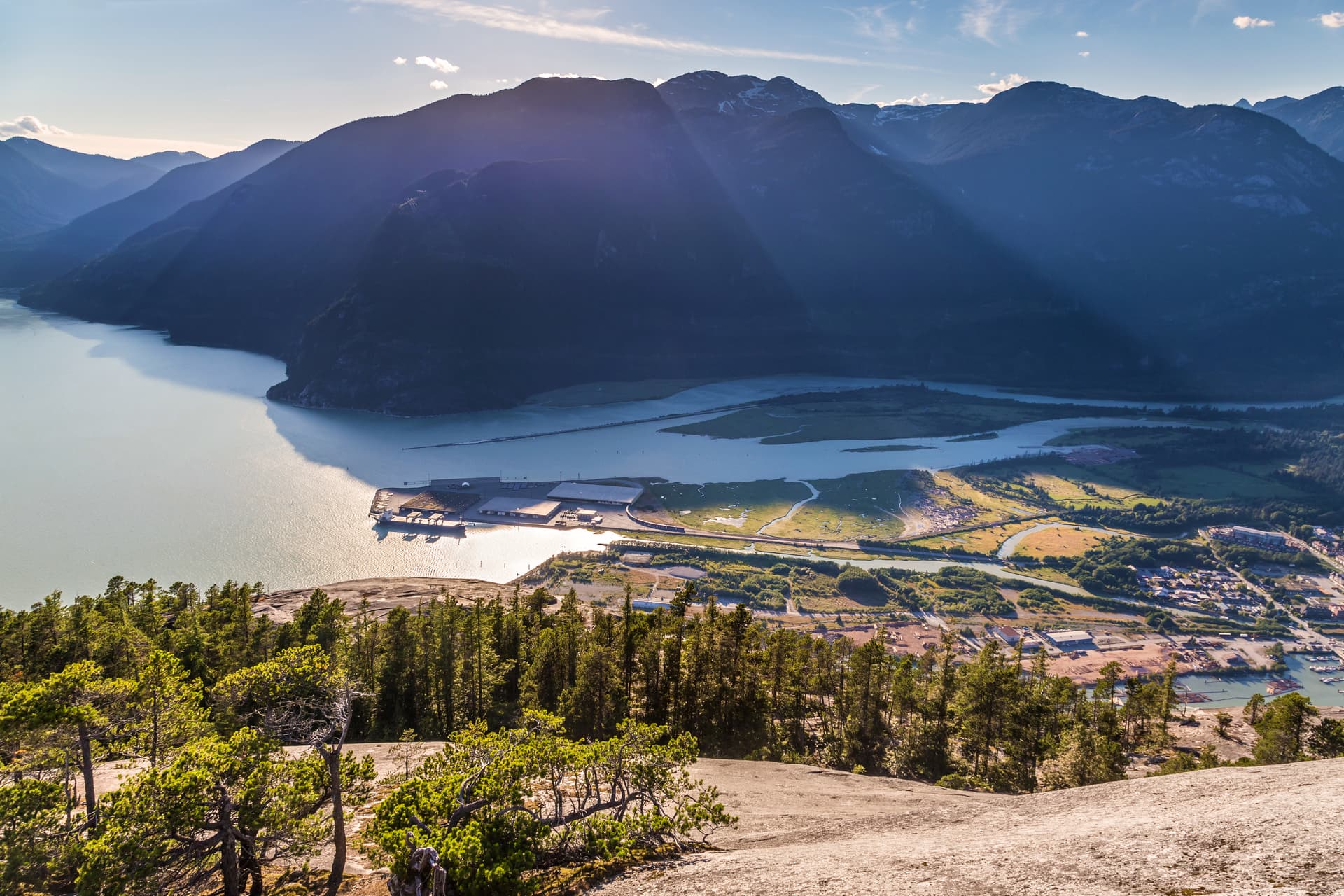 Aerial view of Squamish waterfront, inlet, and forested mountains under bright sky.