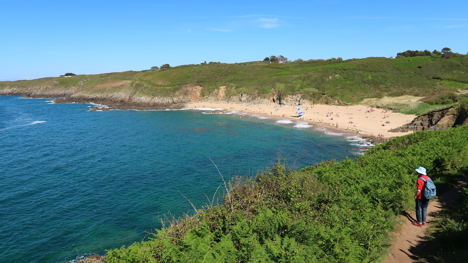 Hiker on path above cove with turquoise water, sandy beach, and small sailboat in Saint-Malo.