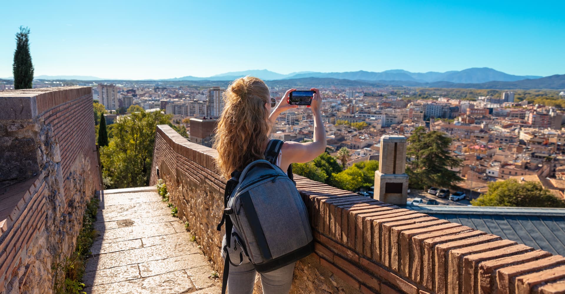 Tourist photographing Girona city view with mountains in the background from stone wall.