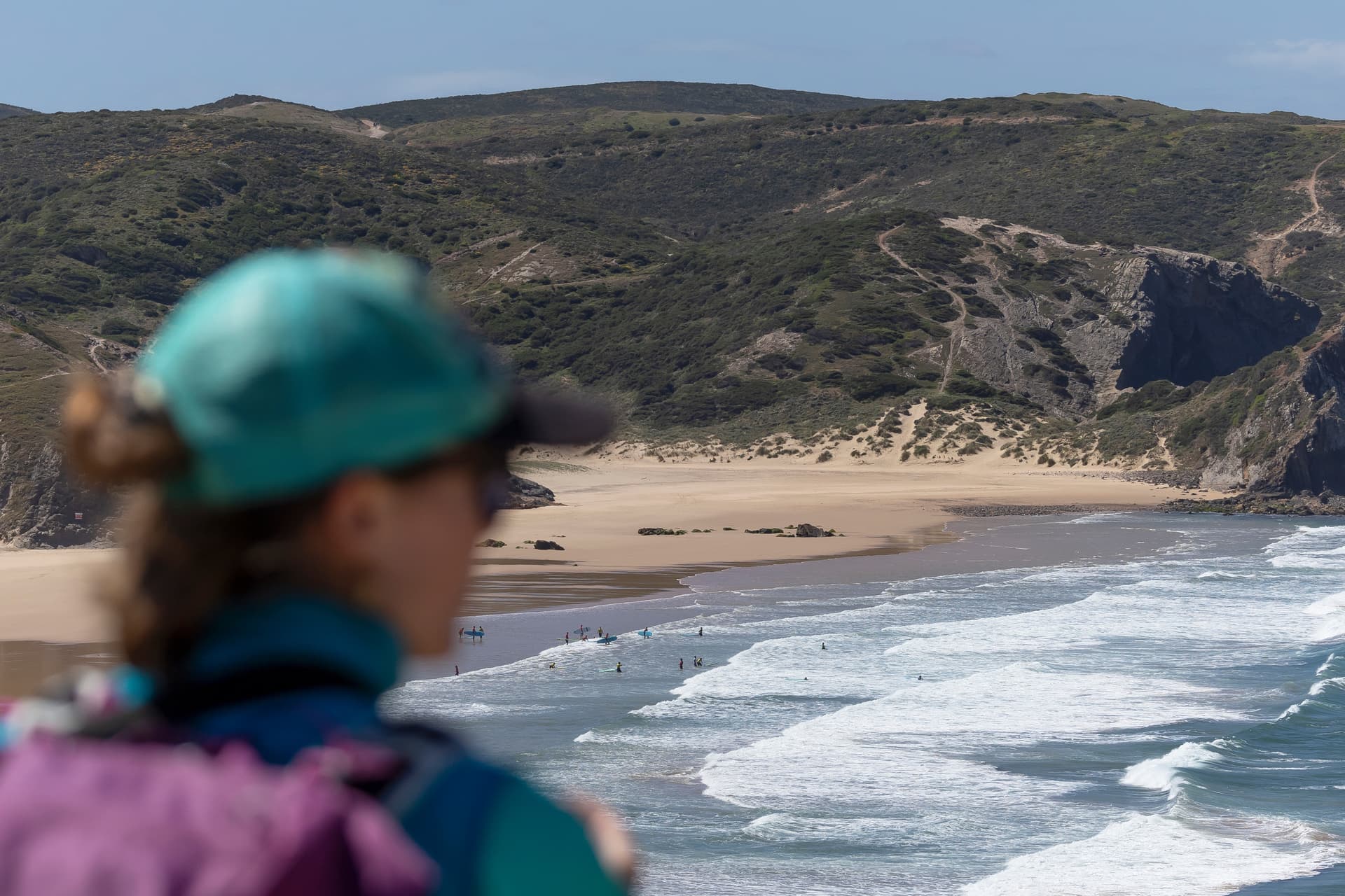 Hiker overlooking surfers on beach with green coastal hills, Rota Vicentina.