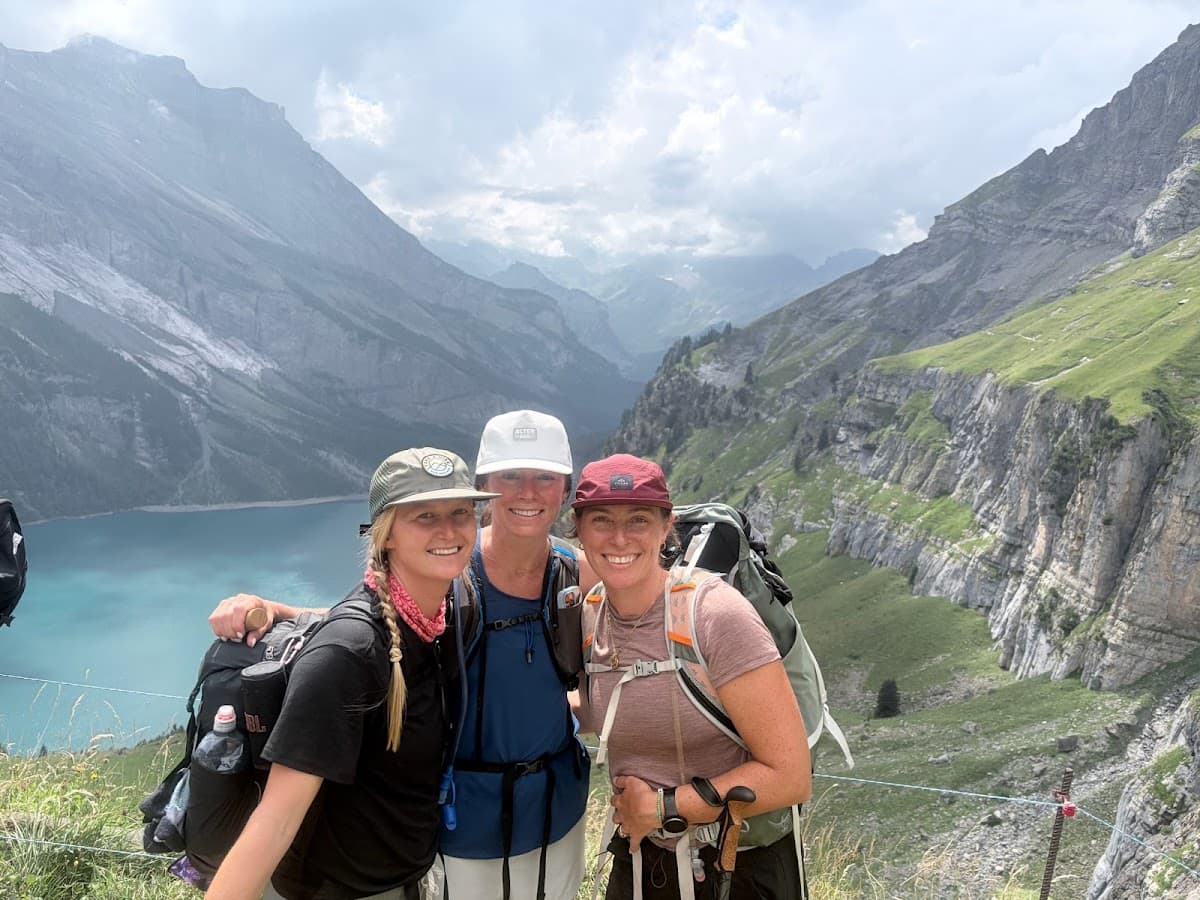 Three women hiking with backpacks near mountain lake and alpine peaks