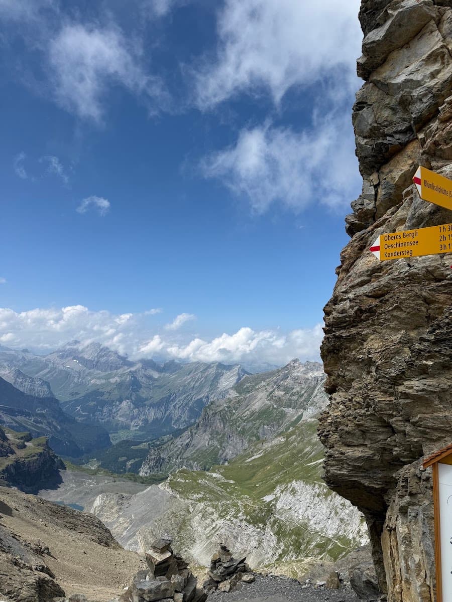 Yellow hiking trail sign mounted on a rocky cliff overlooking alpine mountain peaks and valleys