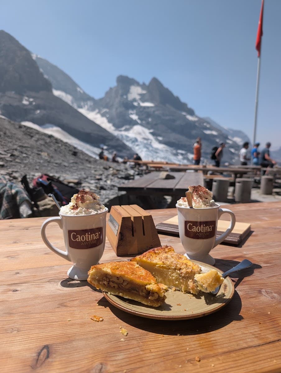 Two mugs of hot chocolate with whipped cream and slices of pastry on an outdoor wooden table with alpine mountain peaks in...