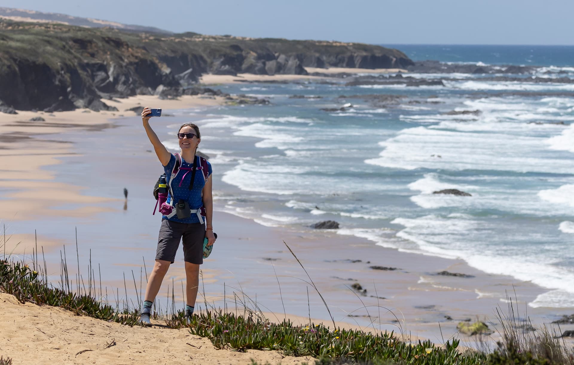 Hiker taking a selfie above a sandy beach with crashing waves and rocky coastline.