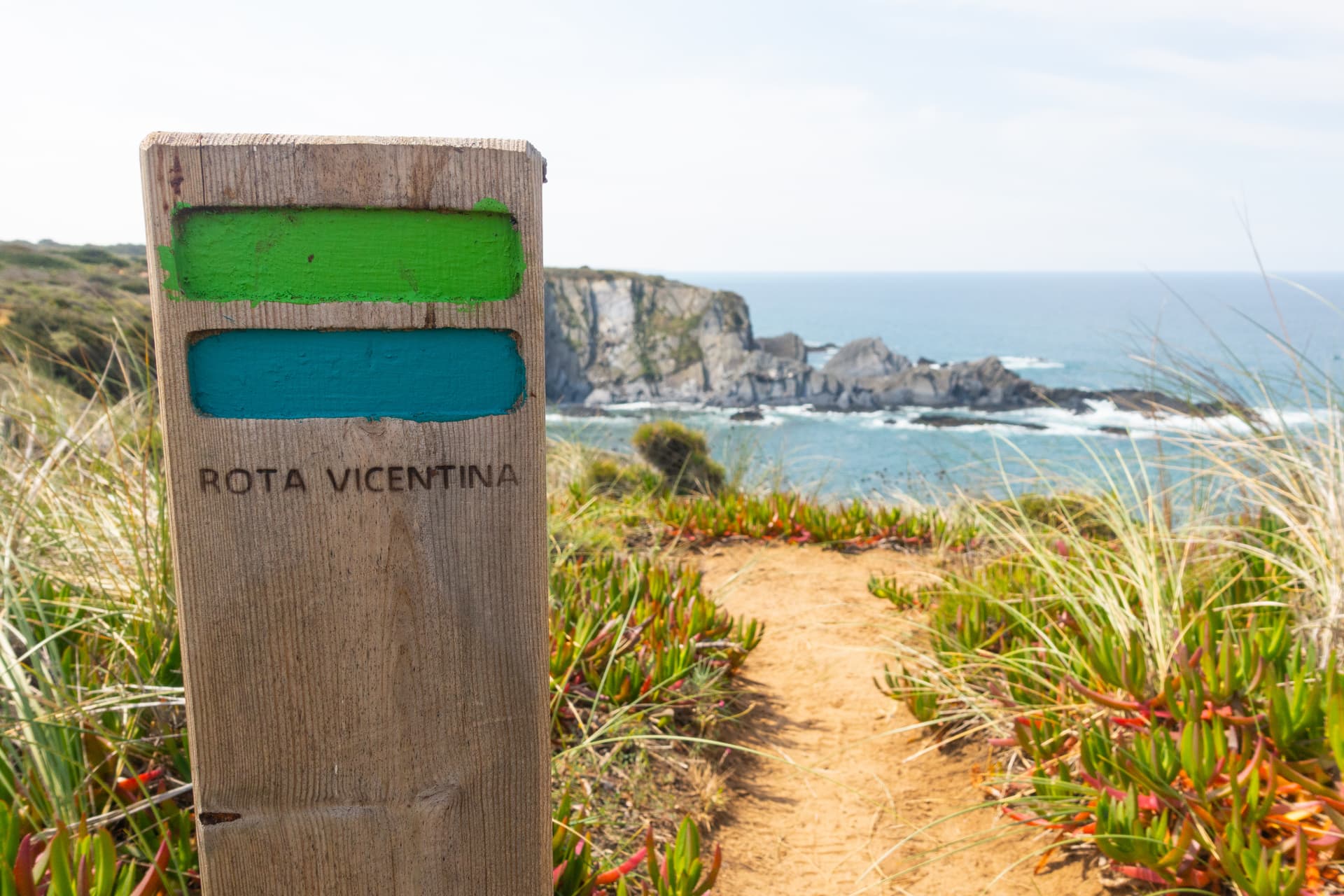 Rota Vicentina trail marker post on dirt path overlooking rocky coastline and blue sea.