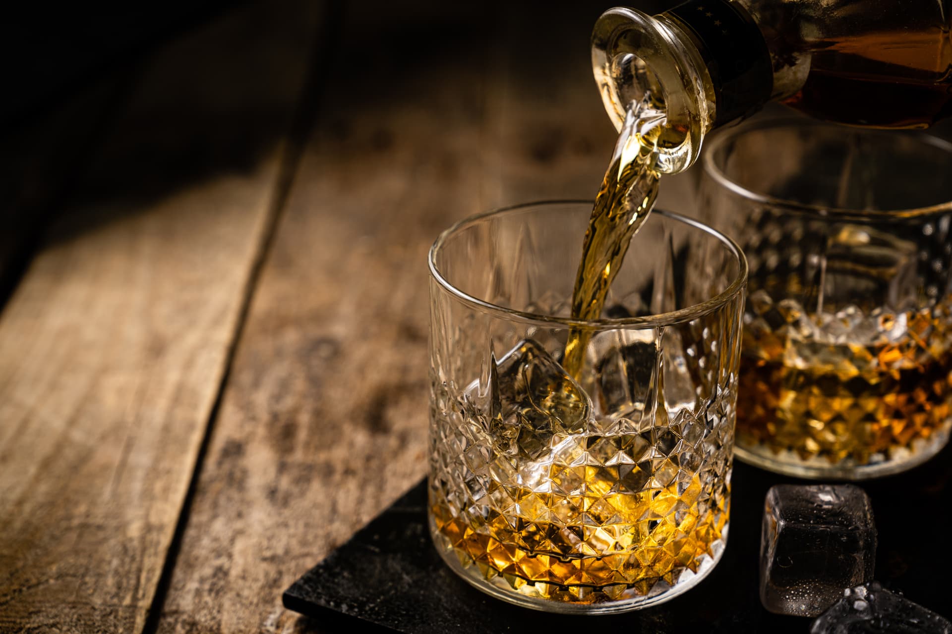 Pouring amber whiskey from a bottle into a crystal glass with ice on a rustic wooden surface.
