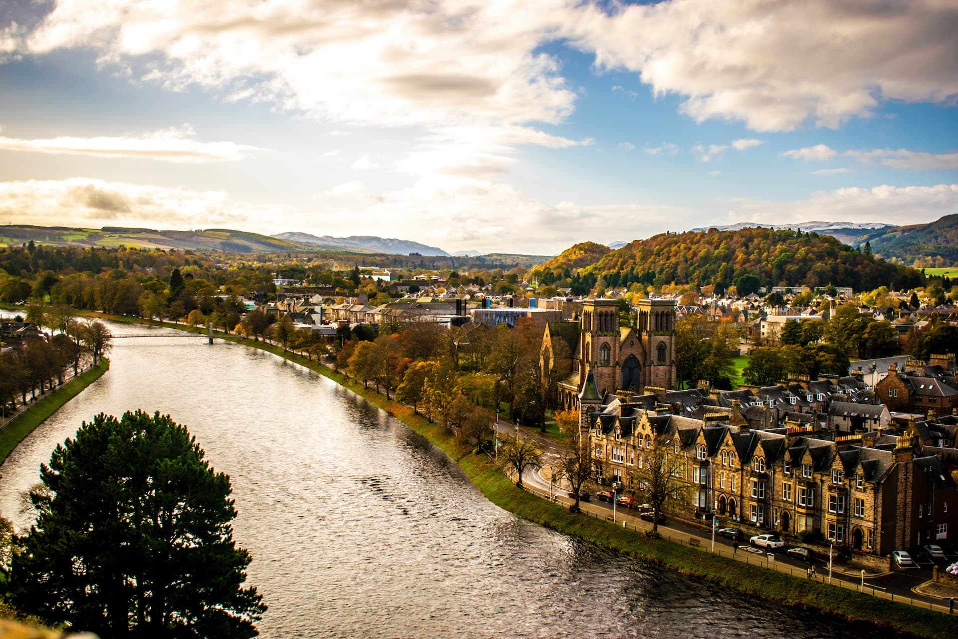 Aerial view of Inverness city with river, autumn trees, and distant hills under cloudy sky.