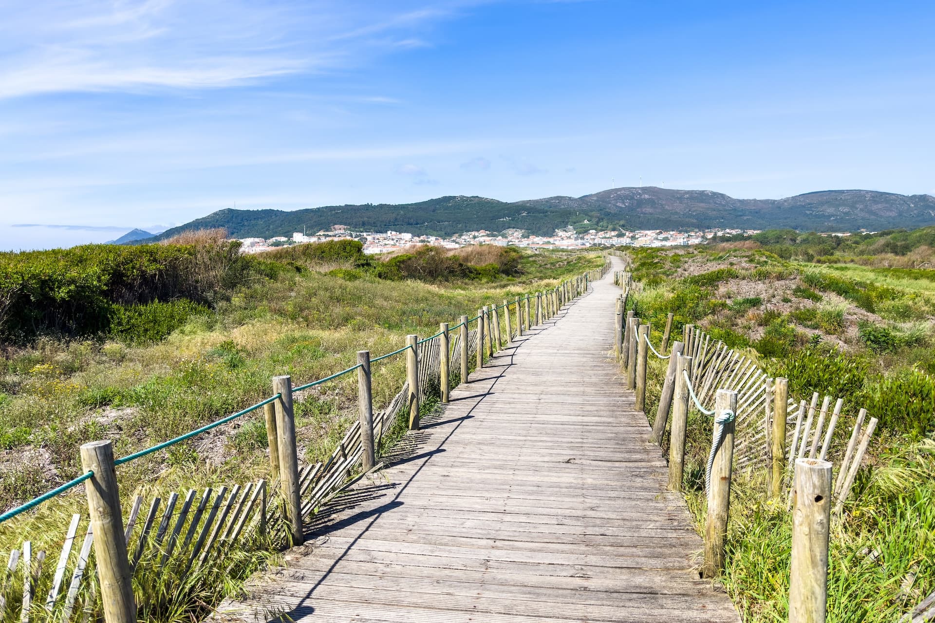 Wooden boardwalk through coastal dunes toward Vila Praia de Âncora town and hills.