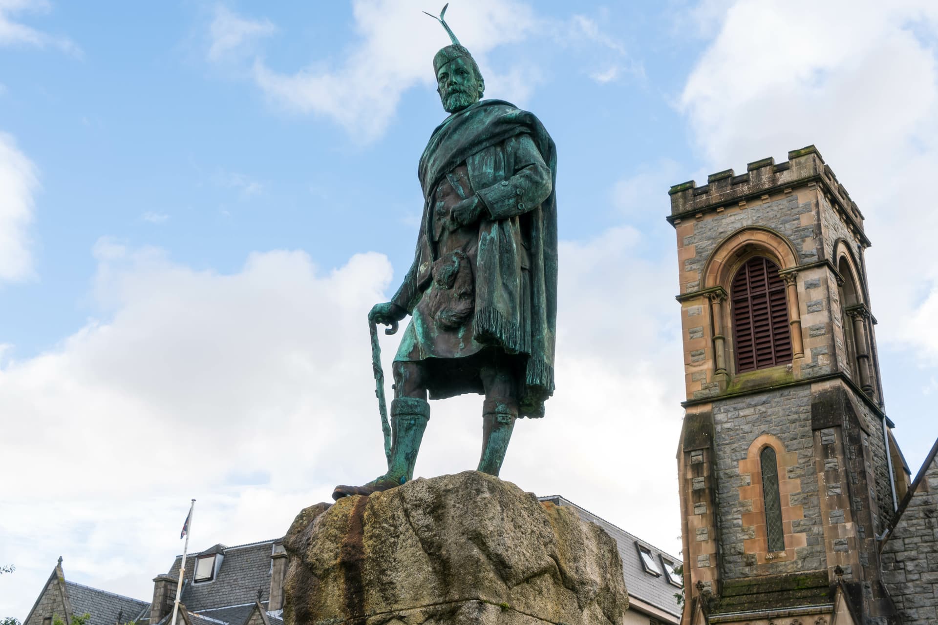Bronze statue of a man in Highland dress on a rock base next to a stone church tower.