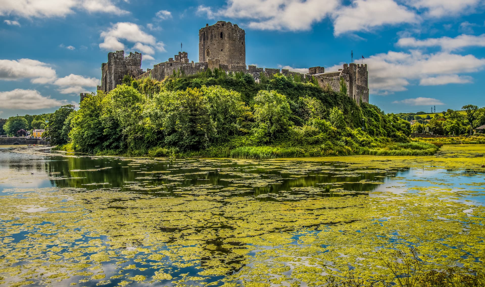 Pembroke Castle on a hill above a moat covered in green algae under a blue sky.