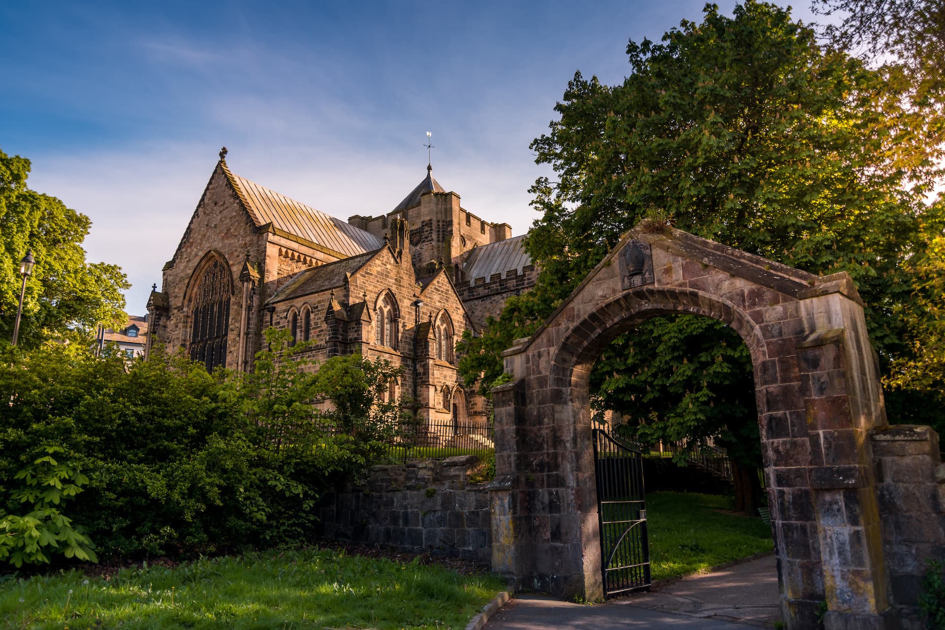 Stone archway entrance to Bangor Cathedral grounds with lush greenery under a blue sky.