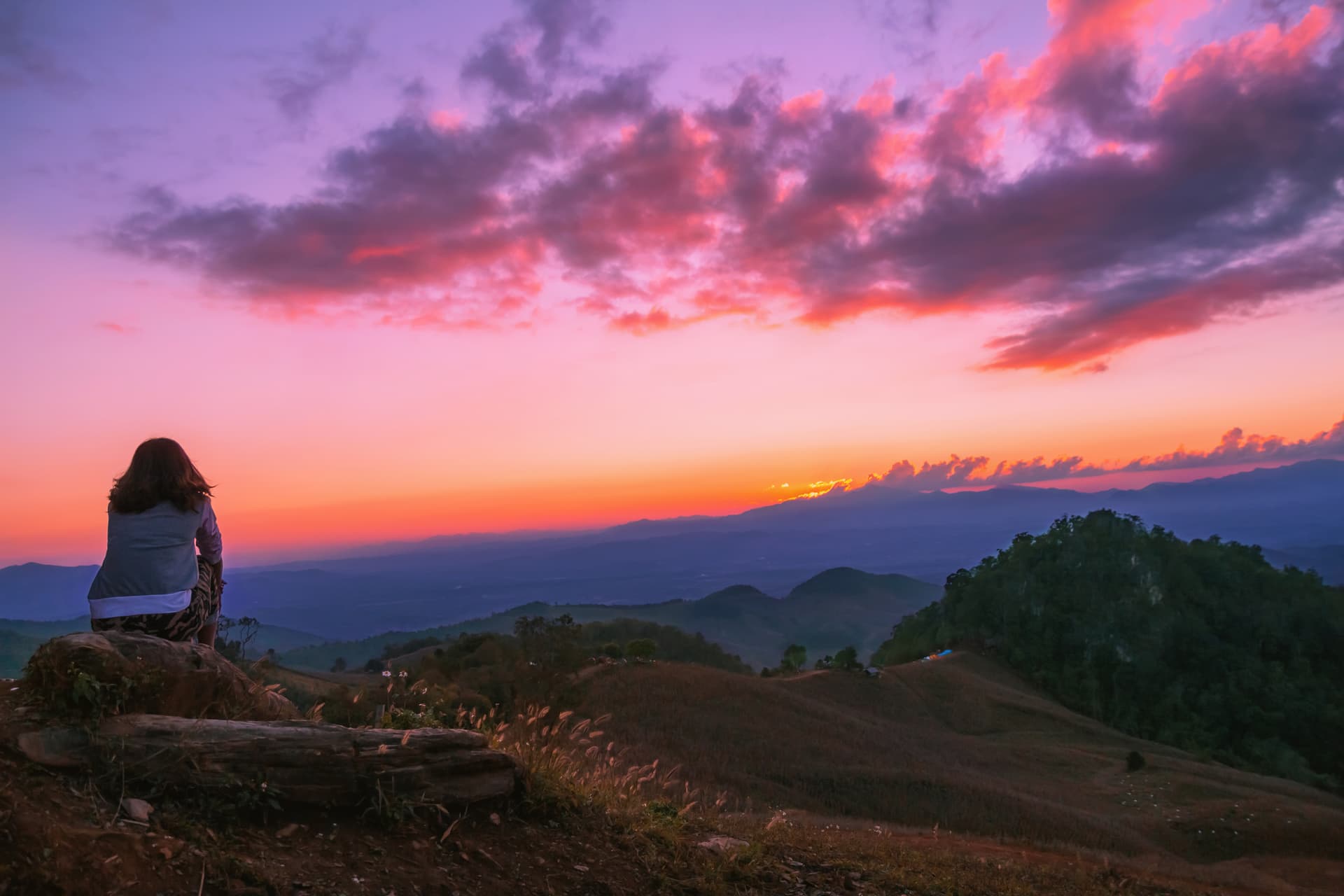 Woman sitting on mountain watching vibrant purple and orange sunset over distant hills