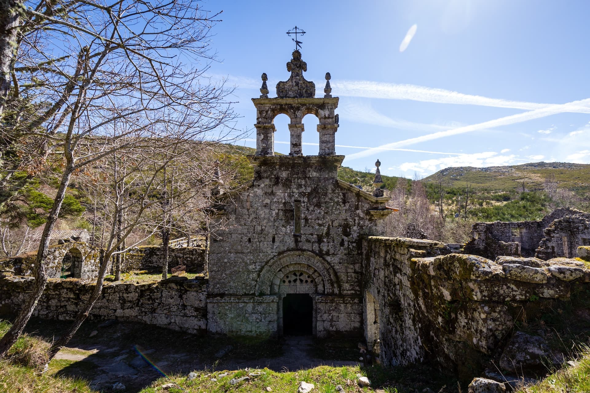Stone ruins of a church with a bell gable surrounded by bare trees and green hills under a blue sky.