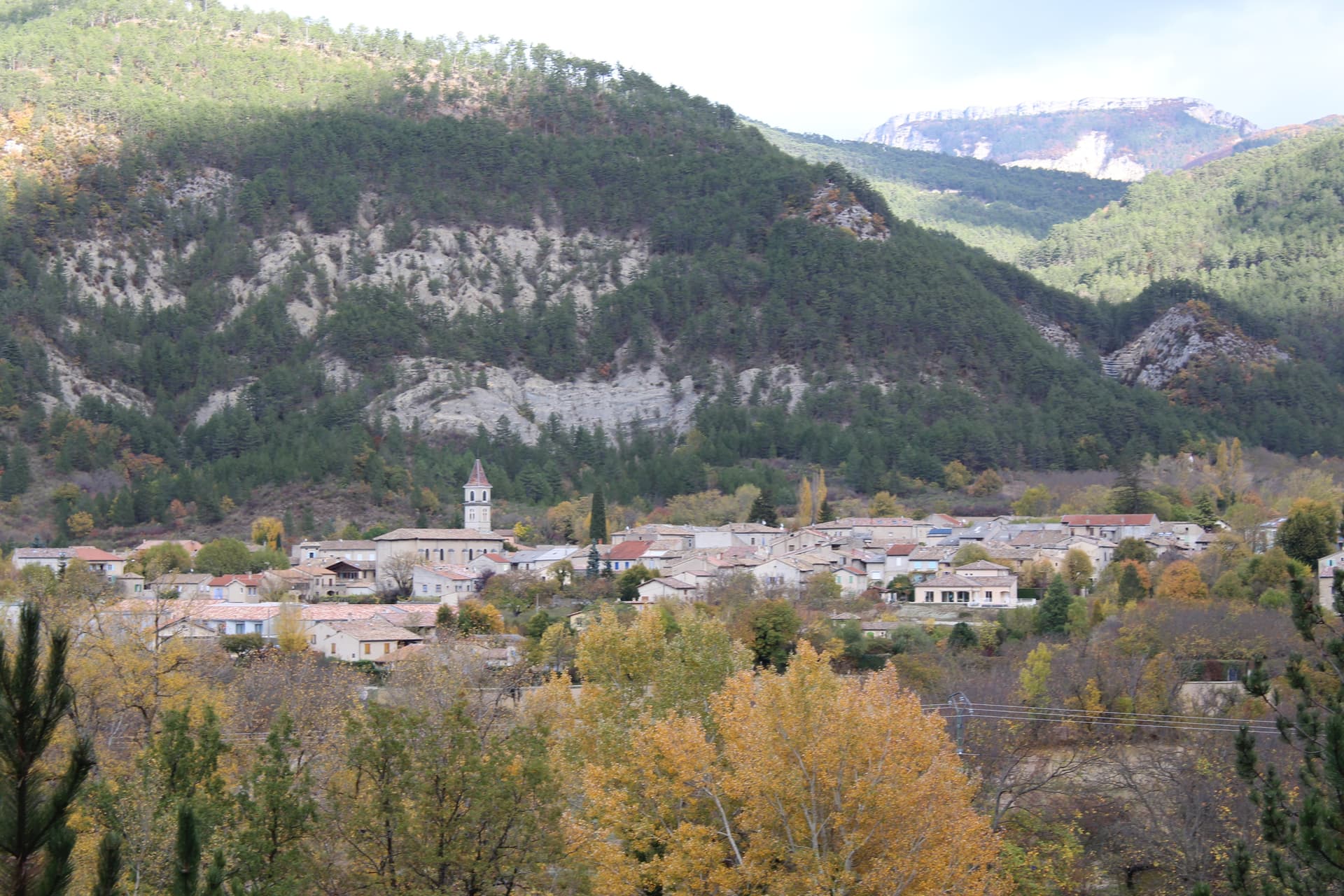 Village nestled in valley with autumn trees below forested mountains, likely in Luc-en-Diois.
