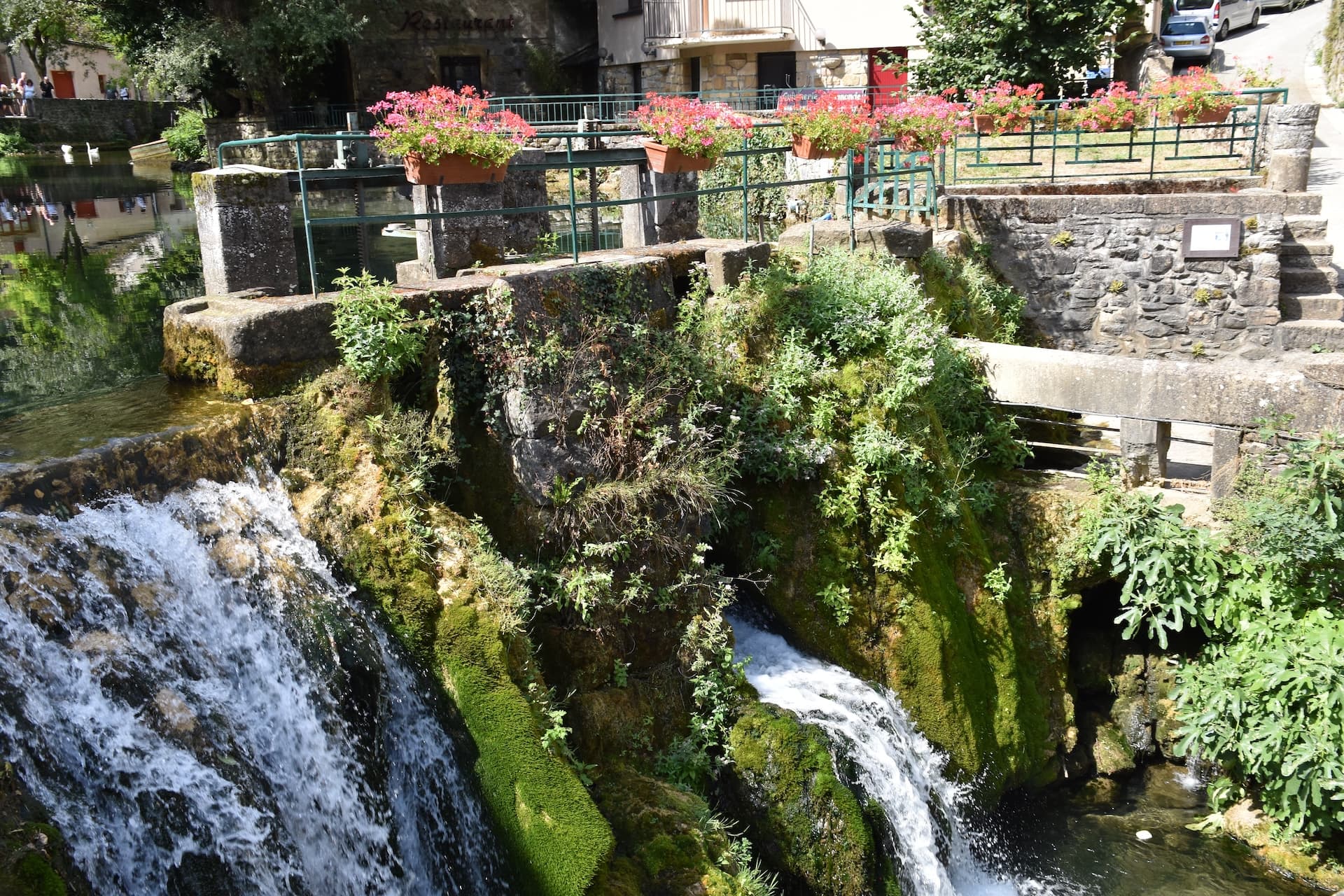 Cascading waterfall over moss-covered rocks near buildings in Florac.