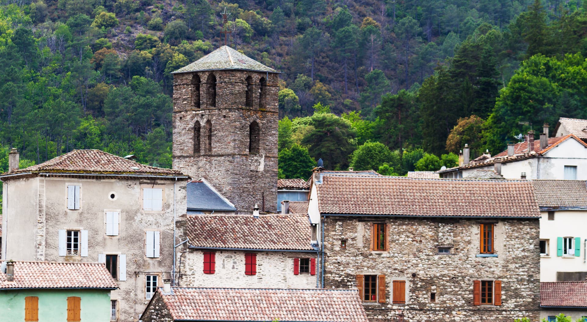 Stone bell tower above village houses with tiled roofs, backed by a dense green forest in Saint-Étienne-Vallée-Française.