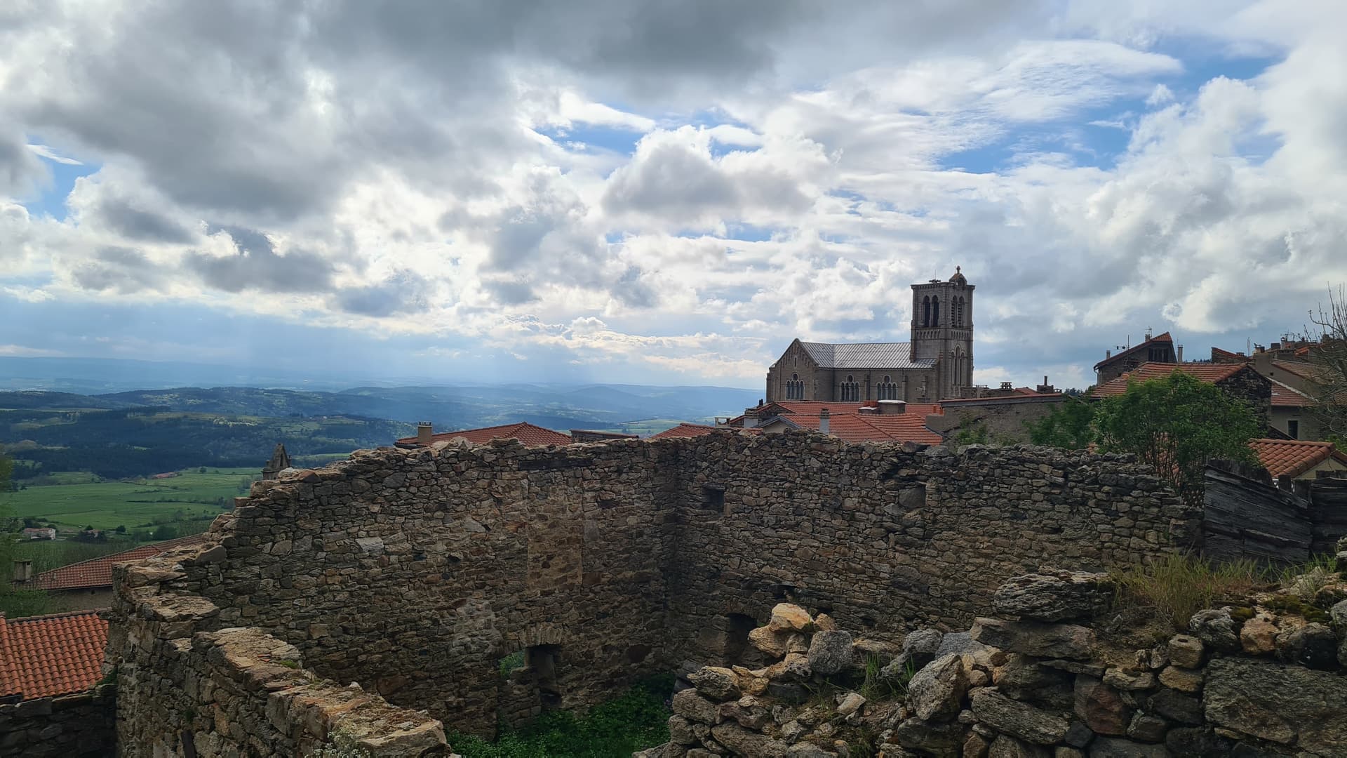 L'église Saint-Clément de Pradelles above stone ruins overlooking rolling green hills under a cloudy sky.